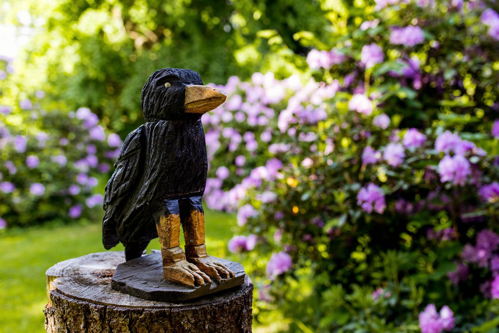Holzfigur eines Vogels auf Baumstumpf im Garten mit lila Blumen.