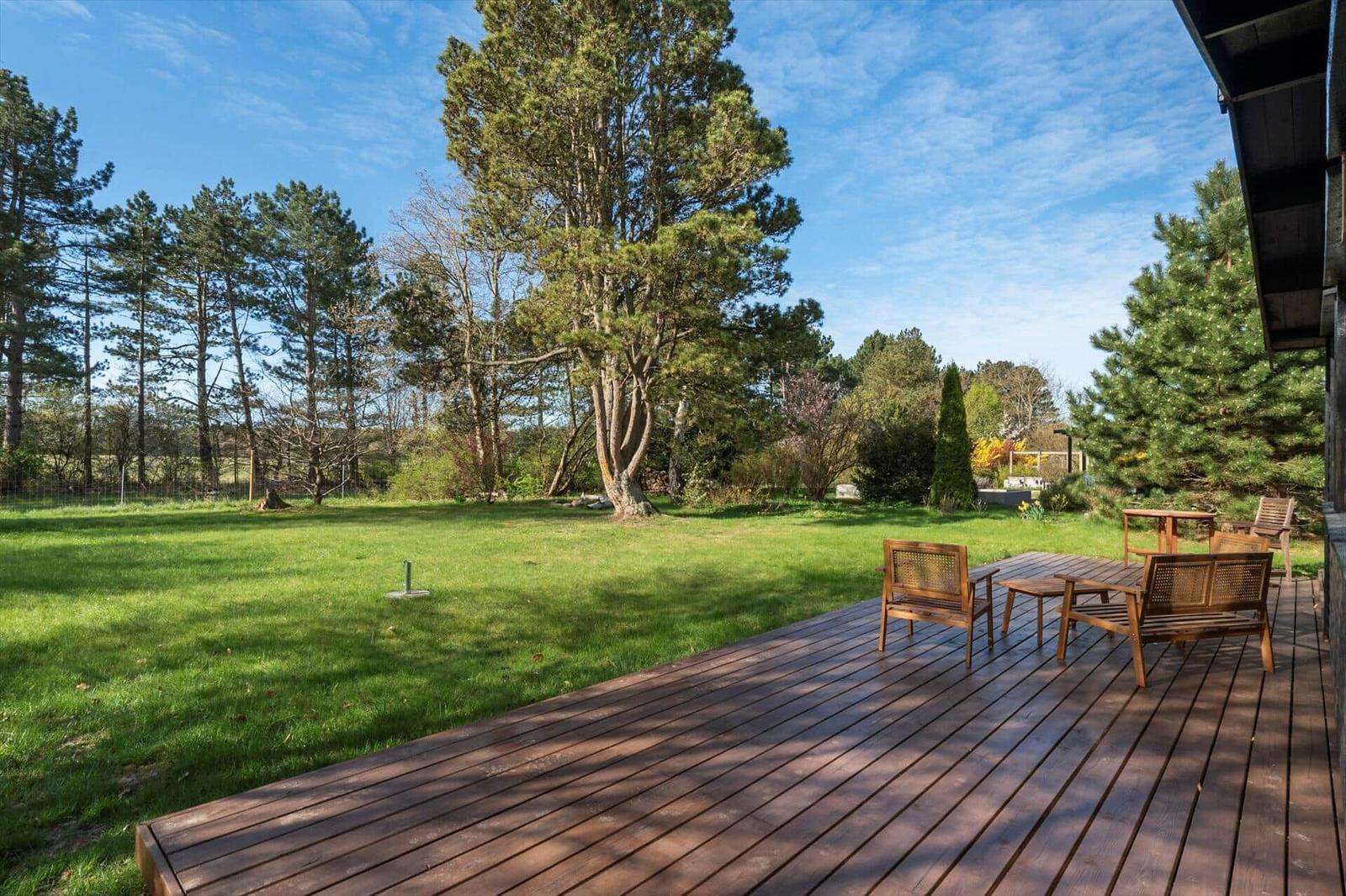Wooden terrace with seating, surrounded by grass and trees under blue sky.