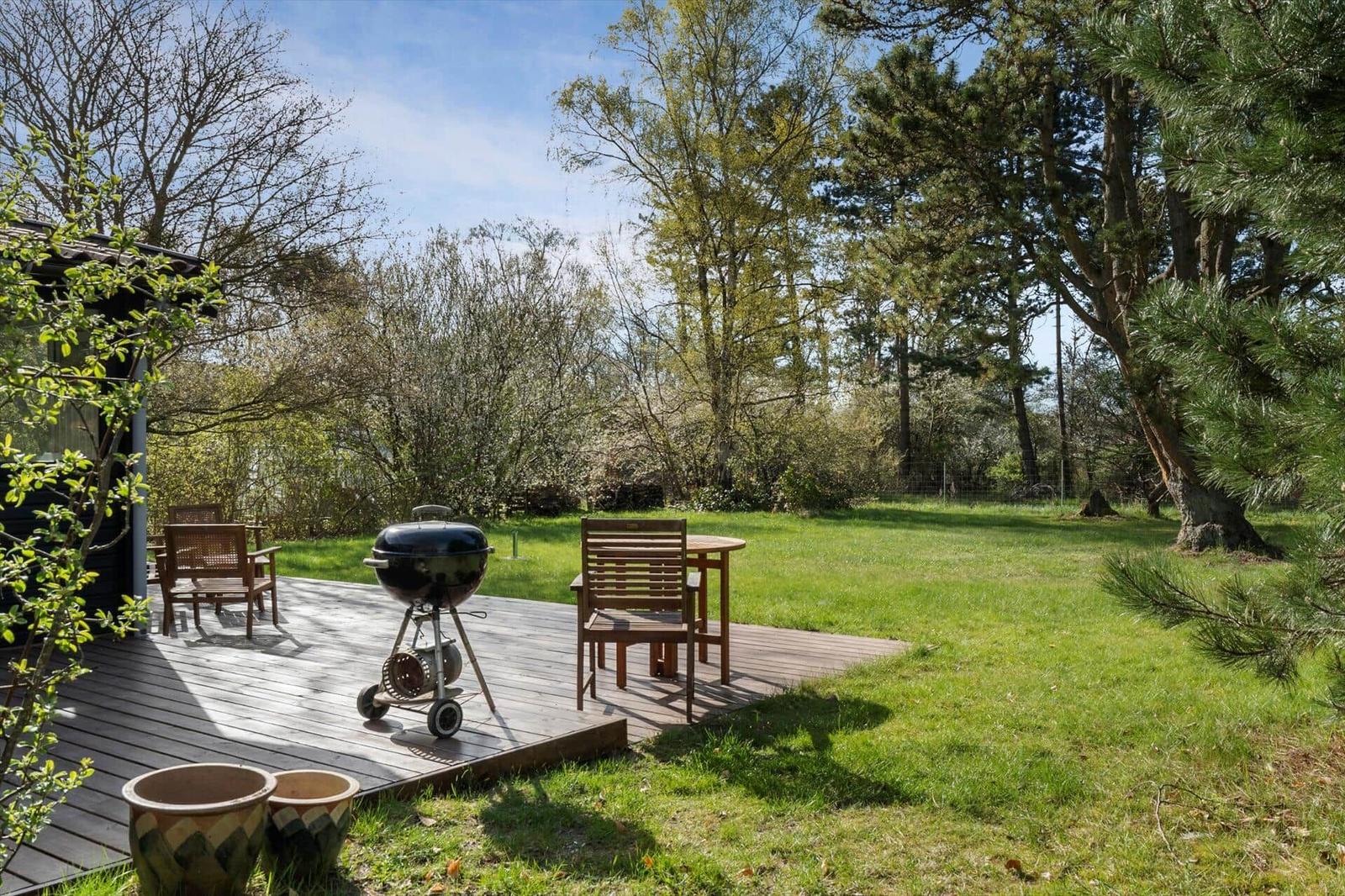 Deck with grill, table and chairs in garden. Lawn and trees in background.