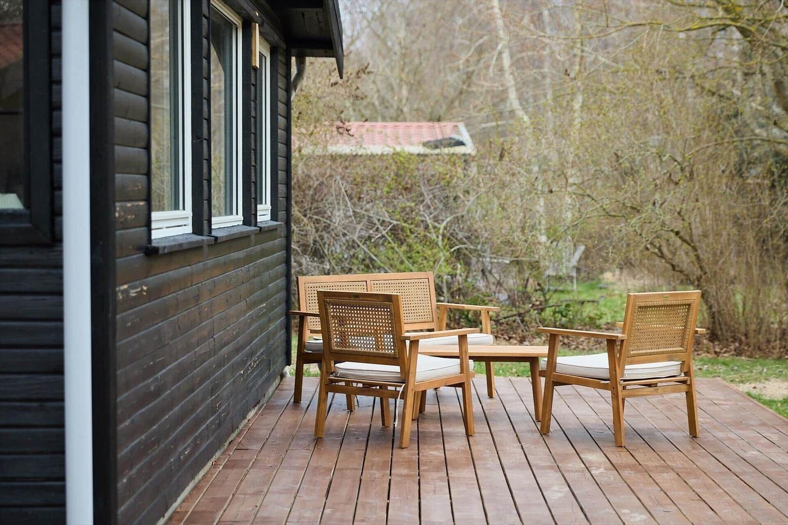 Deck with wooden bench and two chairs in front of a house with dark wood cladding.