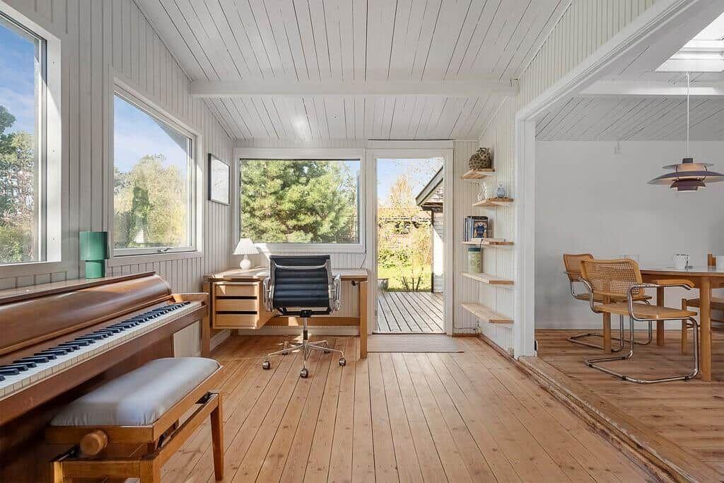 Room with piano, desk, and view of garden. Wooden floor and white walls.