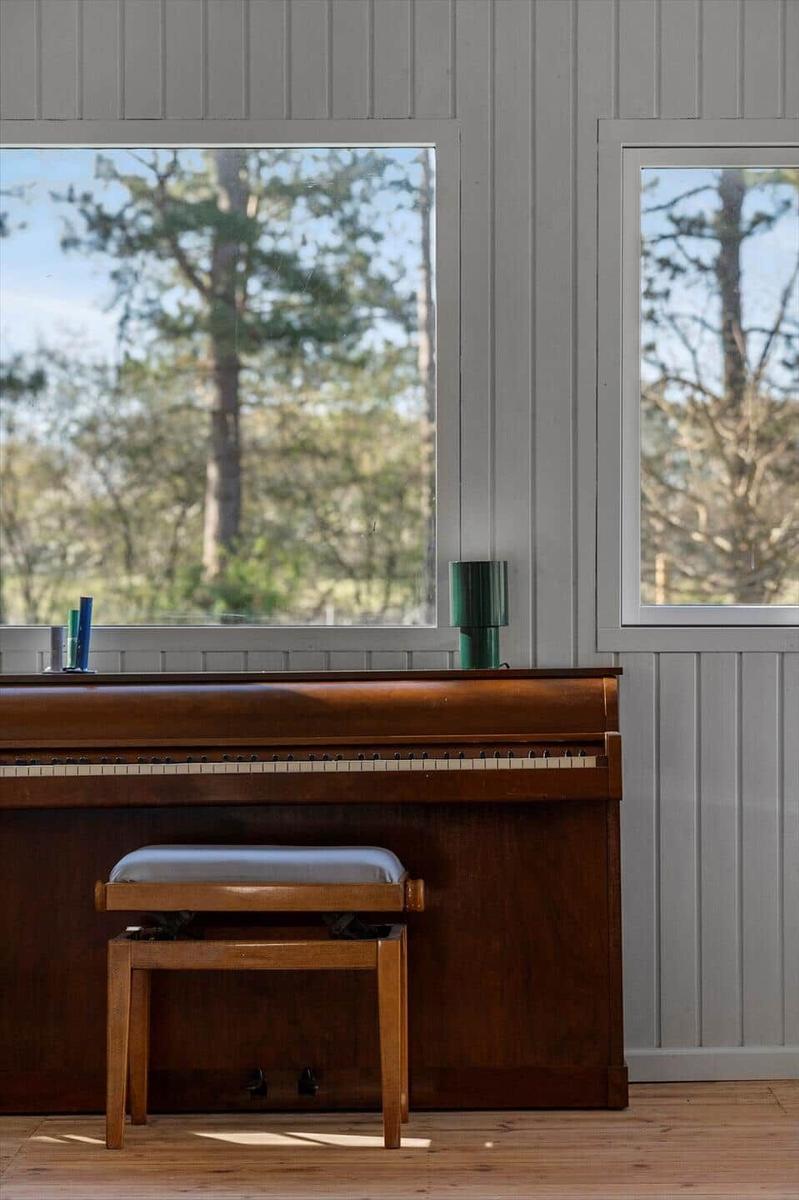 A piano with a stool in front of windows overlooking trees.