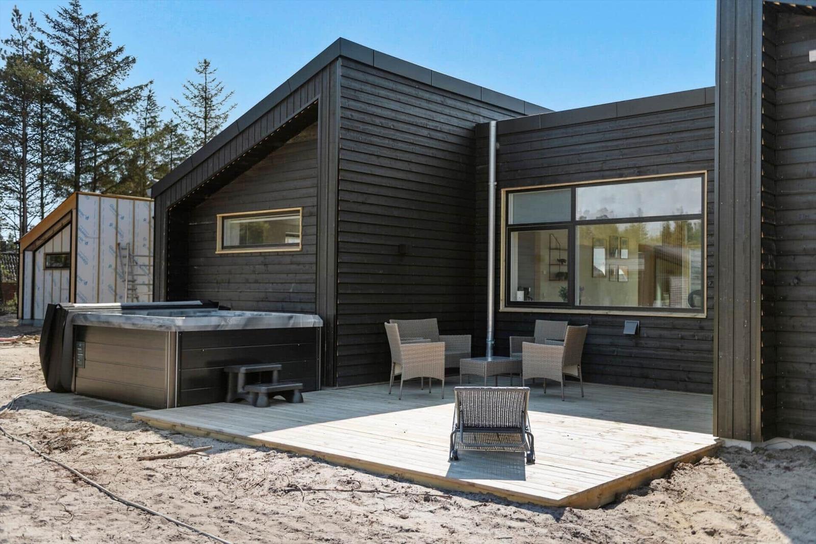 Moderne Holzhütte mit Terrasse, Whirlpool und Gartenmöbeln. Blick auf Wald und klaren Himmel.