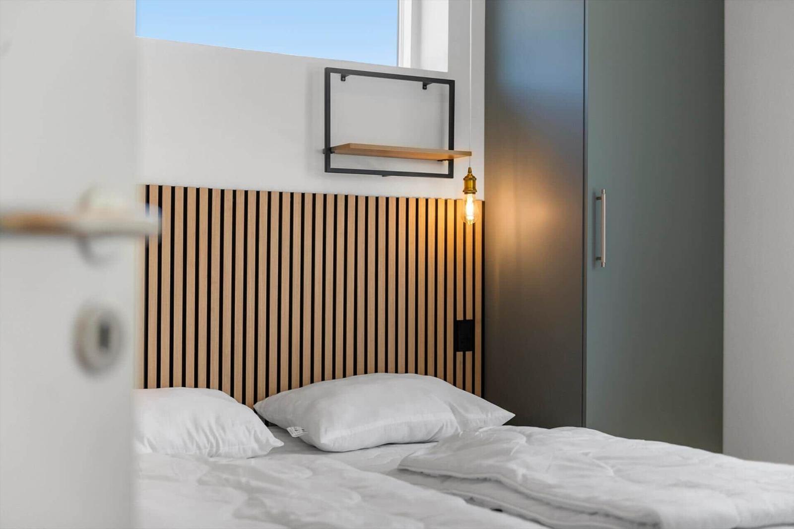 Schlafzimmer mit Bett, Holzkopfteil und Wandregal. Helle Wände und modernes Licht.