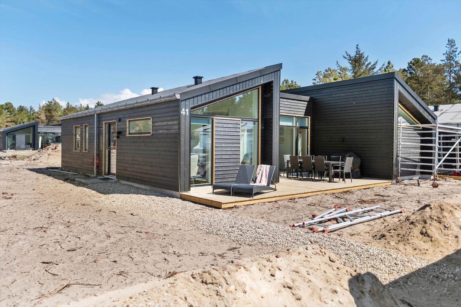 Moderne Holzhütte mit Terrasse, Liegen und Tisch. Im Hintergrund Wald. Baustelle im Vordergrund.
