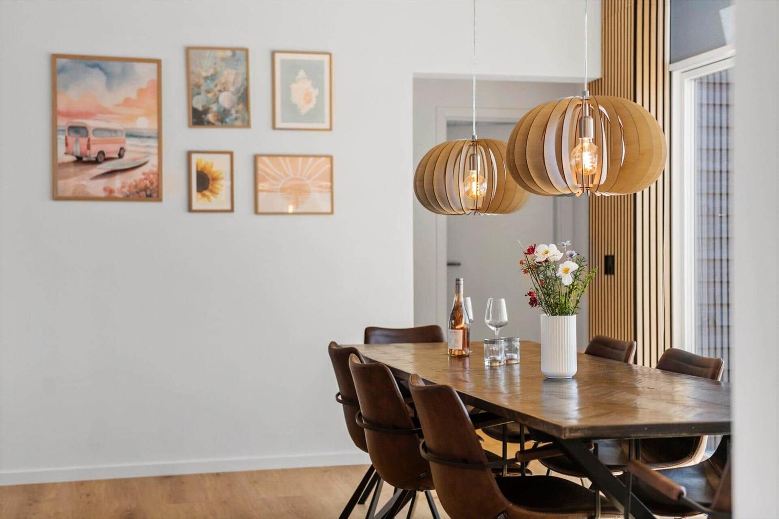 Esszimmer mit Holztisch, Stühlen und hängenden Lampen. Wand mit Bildern und Fenster mit Jalousie.
