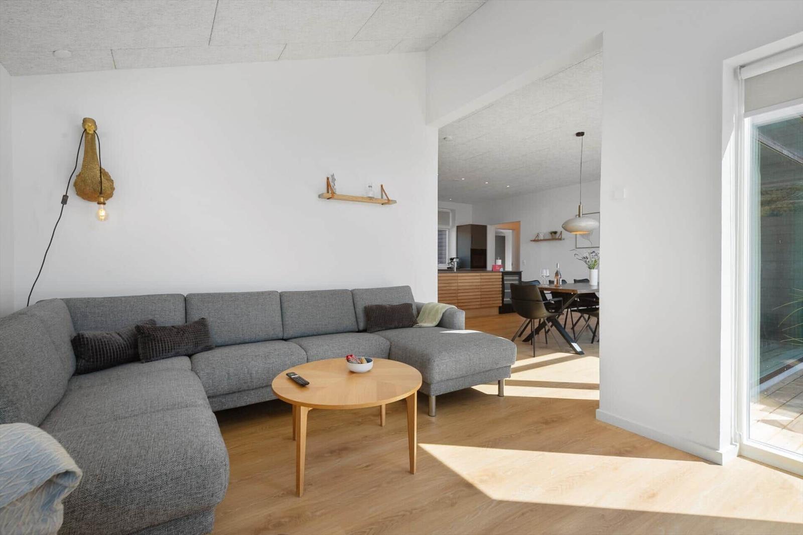 Ein Wohnzimmer mit grauem Sofa, runden Tisch und Holzboden. Durchgang zur Küche und Terrasse.