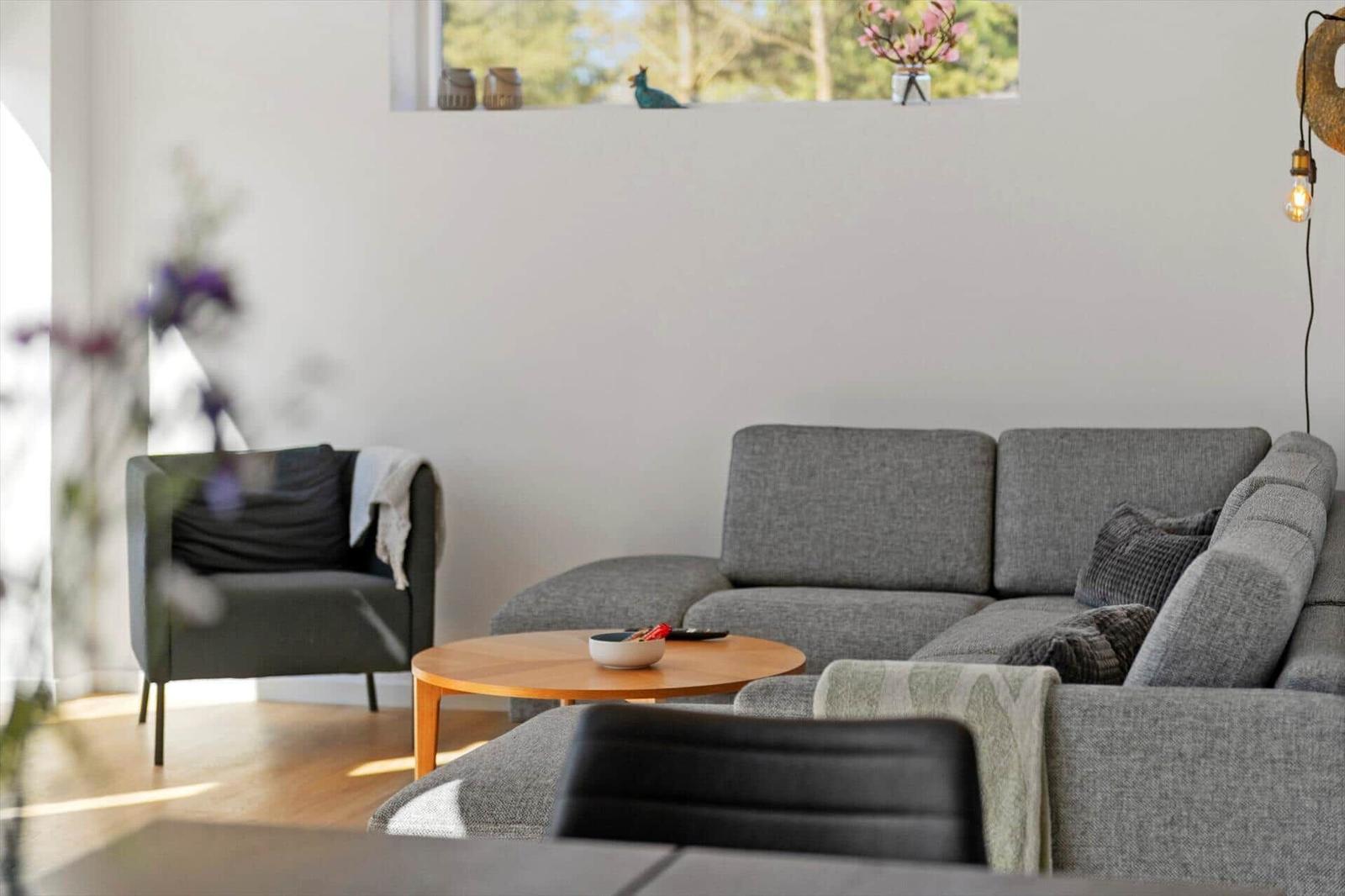 Ein Wohnzimmer mit grauem Sofa, Holztisch und Sessel. An der Wand hängt ein Bild mit Blumen.