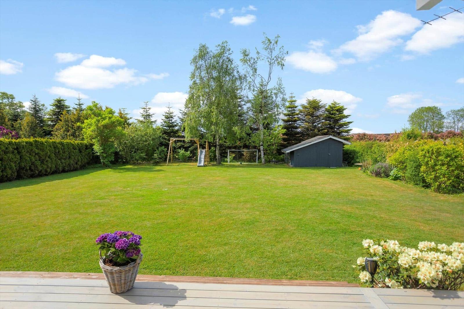 Large green garden with playground, shed, and flower beds. Wooden terrace in the foreground.