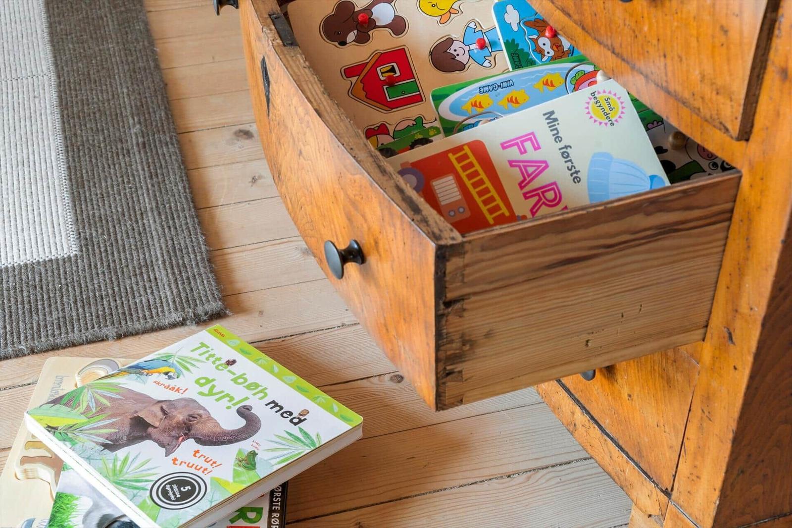 Holzschrank mit geöffneter Schublade, die Kinderbücher und Puzzles enthält, auf Holzboden mit Teppich.