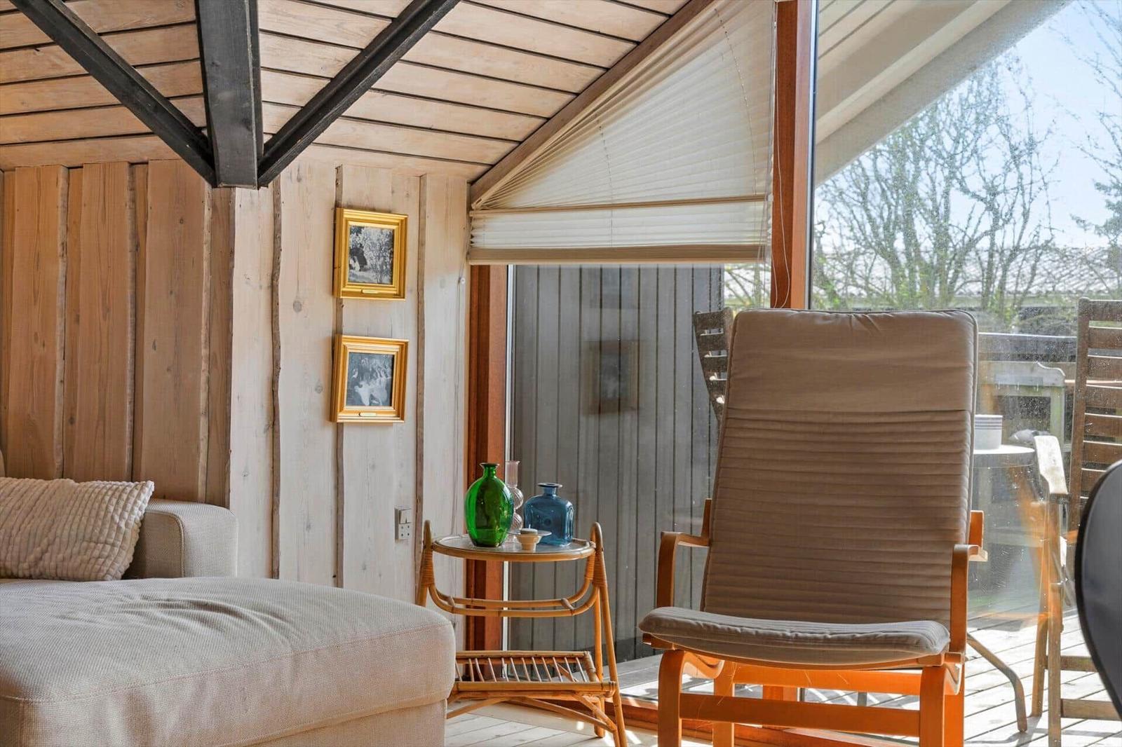 Ein Raum mit Holzwand und -decke, einem Sessel und einem Sofa. Durch das Fenster ist eine Terrasse sichtbar.