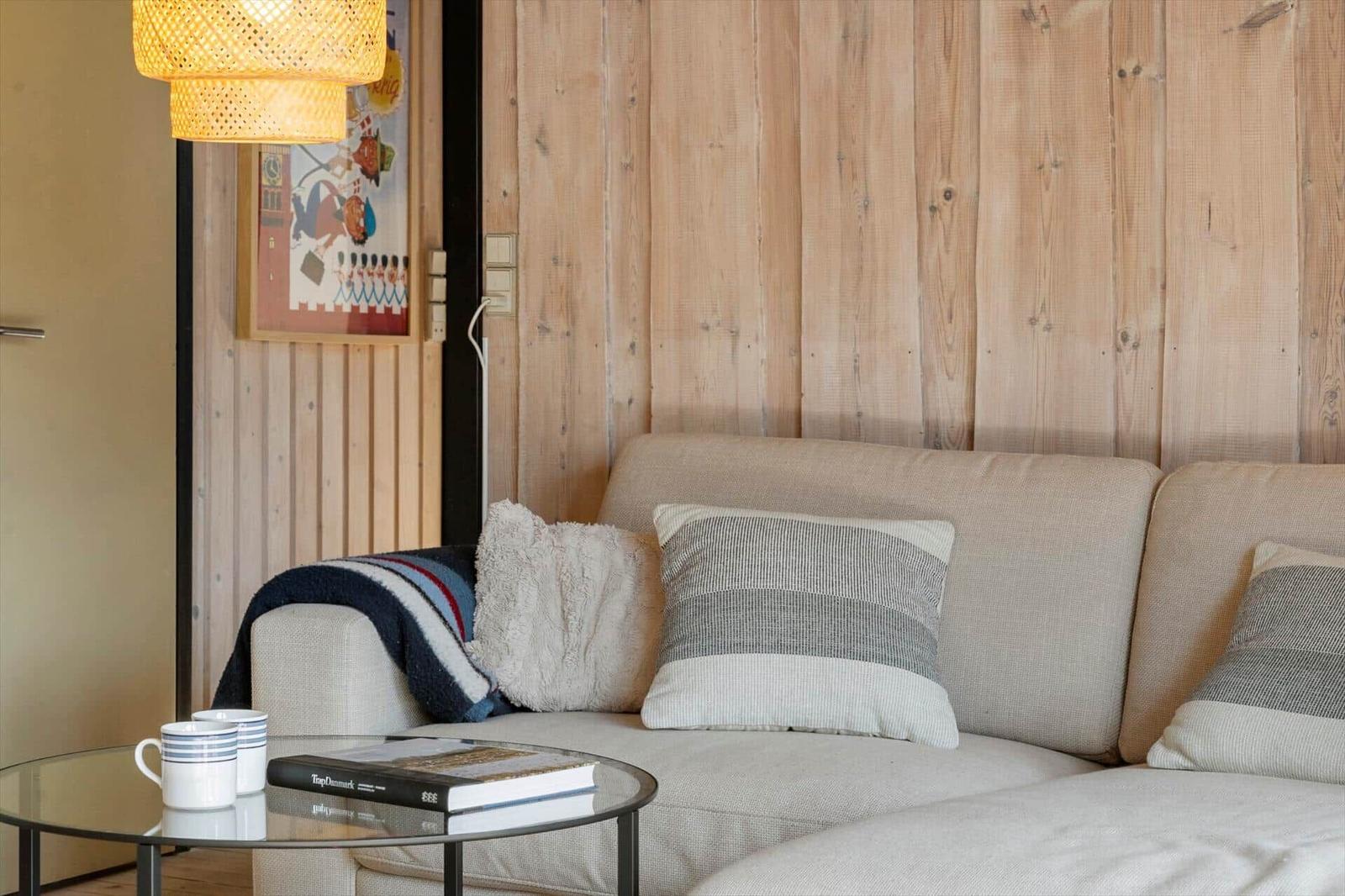 Ein Sofa mit Kissen und Decke vor einer Holzwand. Auf einem Tisch stehen Tassen und ein Buch.