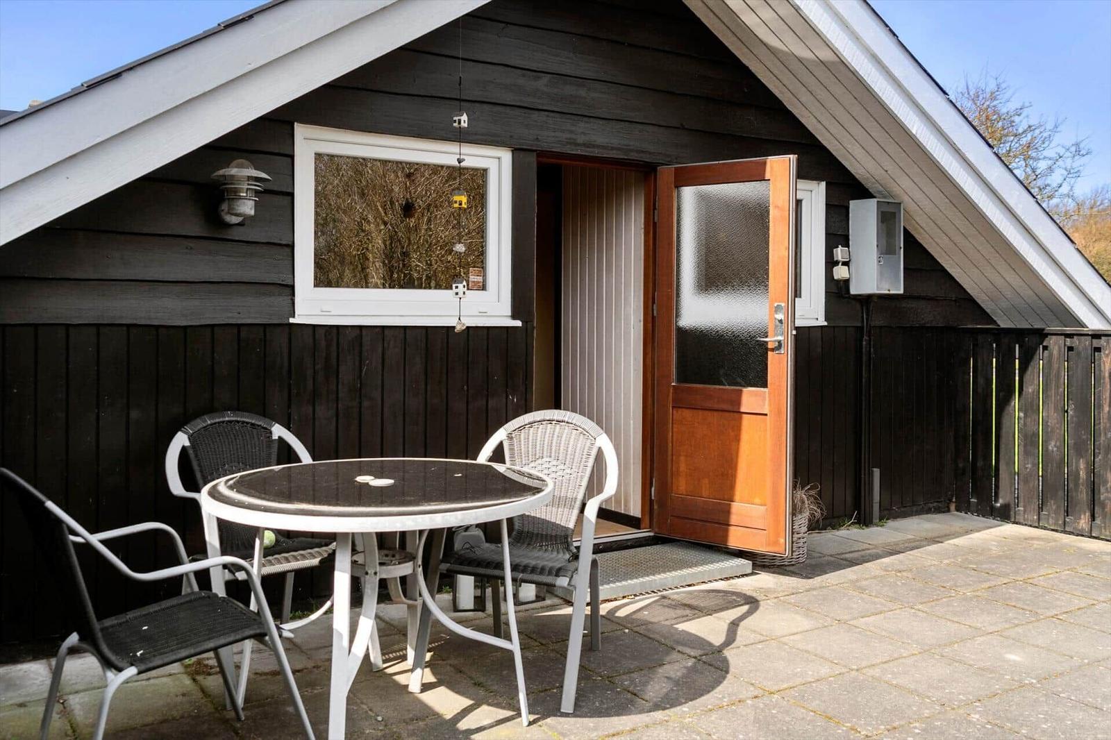 Haus mit Terrasse, Tisch und Stühlen. Tür offen. Holzwand und Dach.