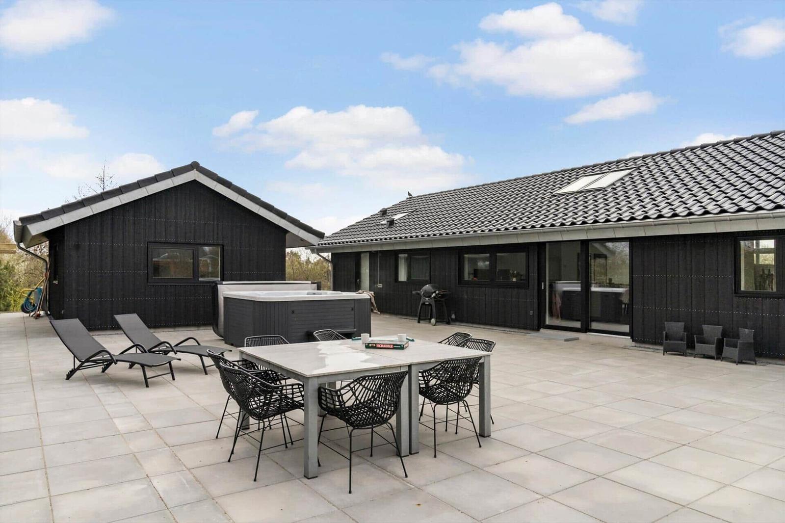 Großer Außenbereich mit Terrasse, Liegestühlen, Esstisch und Whirlpool. Schwarze Holzfassade und Dachziegel.