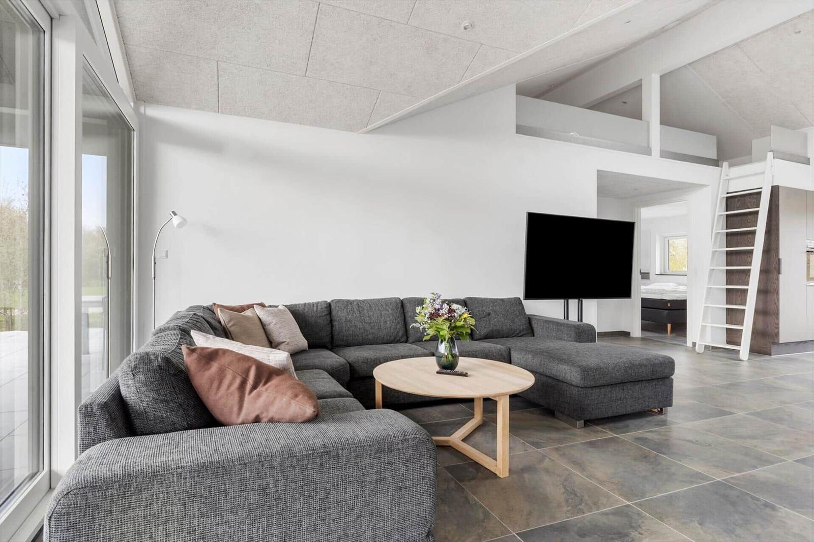 Wohnbereich mit grauem Sofa, runden Tisch und Fernseher. Hintergrund zeigt Schlafbereich mit Leiter.