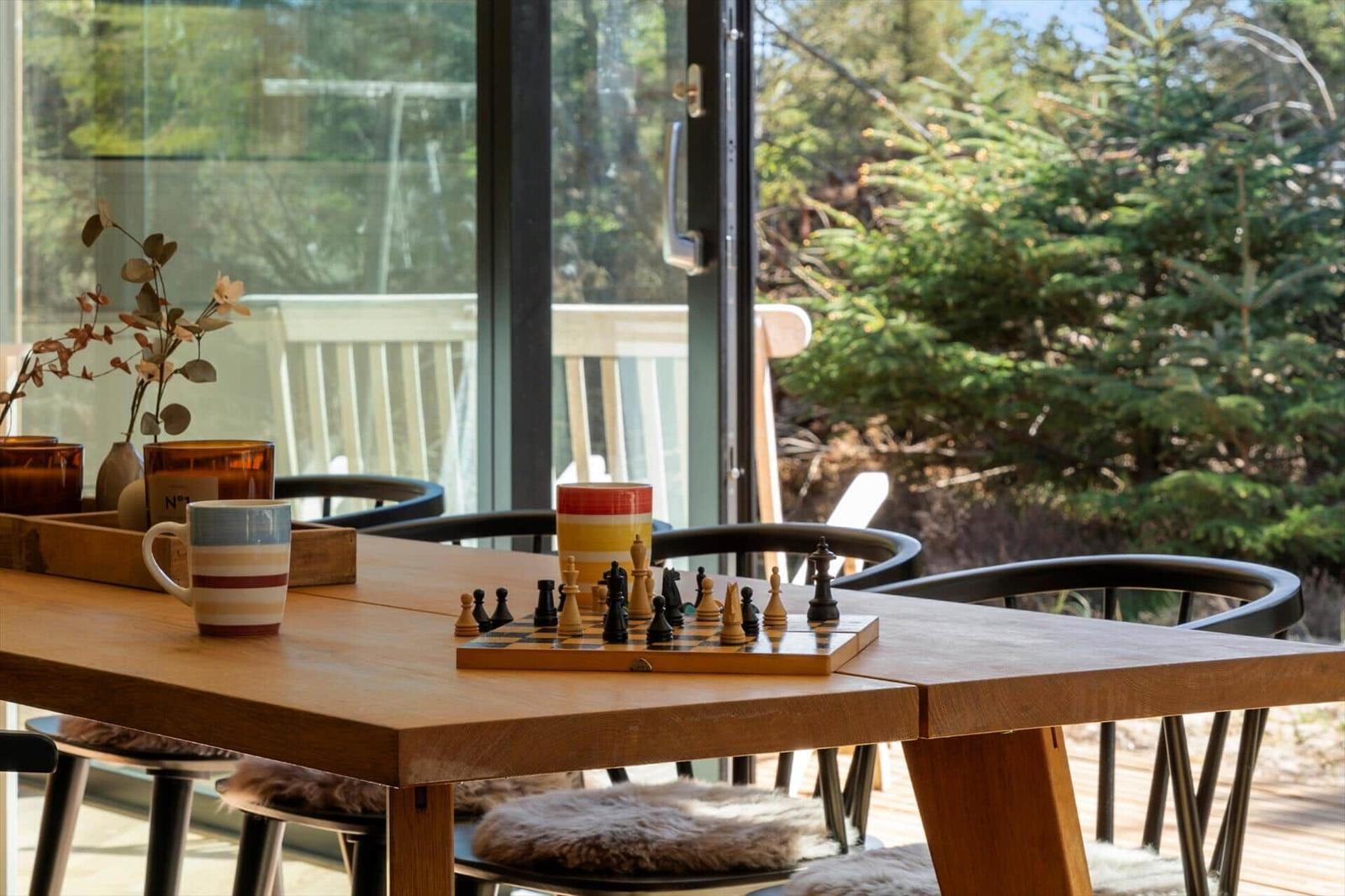 Wooden table with chessboard and mugs in front of large window with forest view.