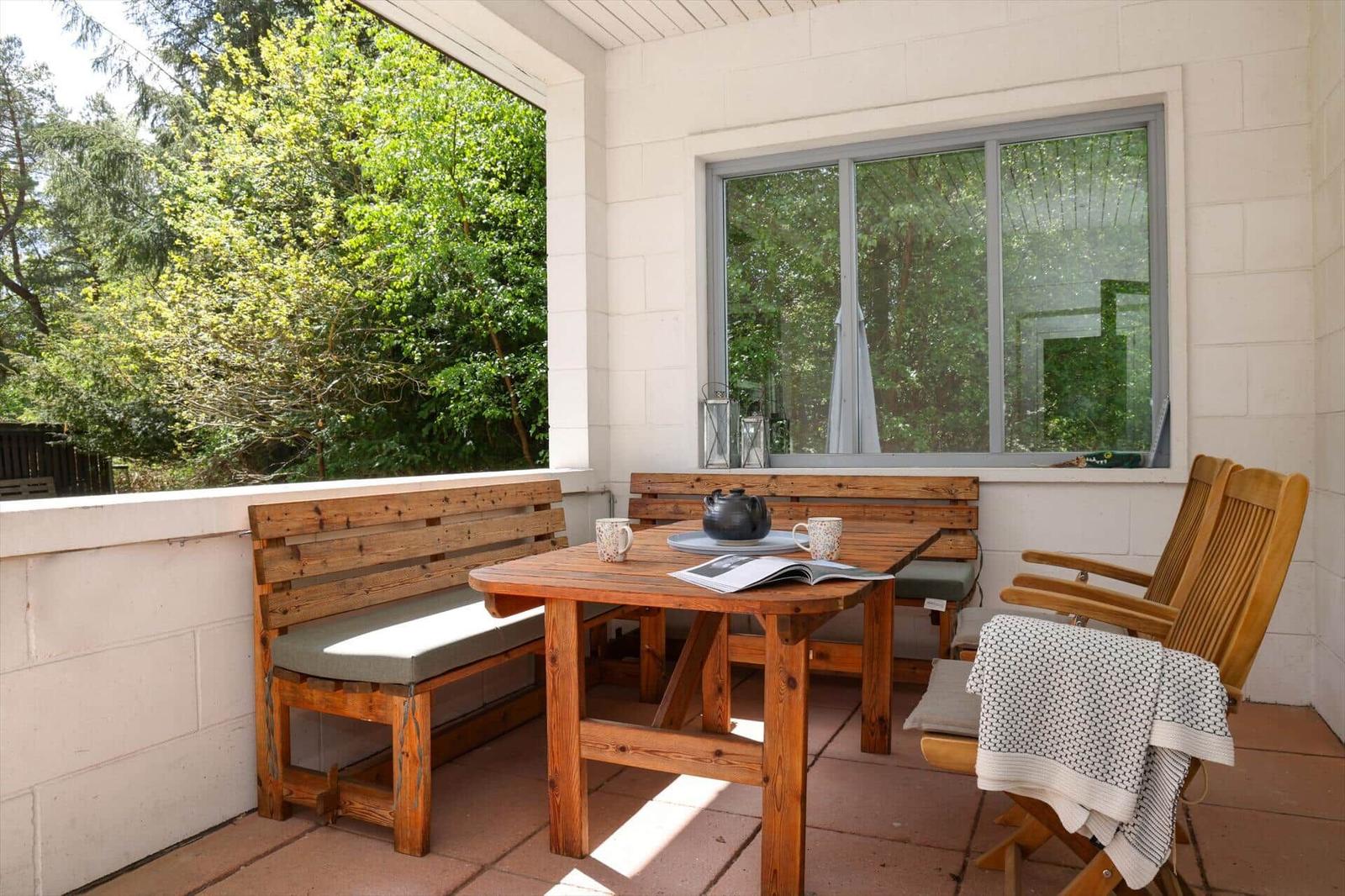 Terrasse mit Holztisch, Bänken und Stühlen. Im Hintergrund sind Bäume sichtbar.