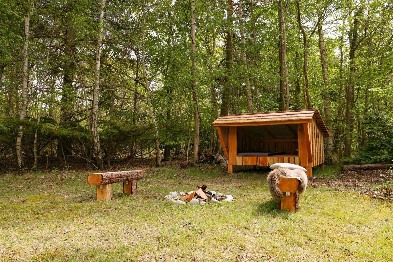 Holzkonstruktion mit Bett, Feuerstelle und Bänken im Wald.