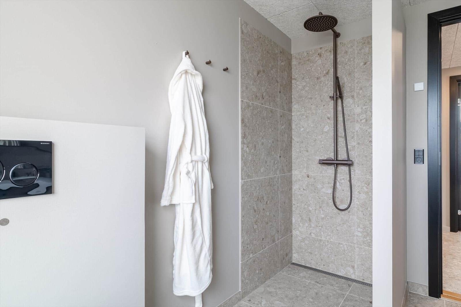 Dusche mit Steinfliesen und Duschbrause. Bademantel hängt an Wand.