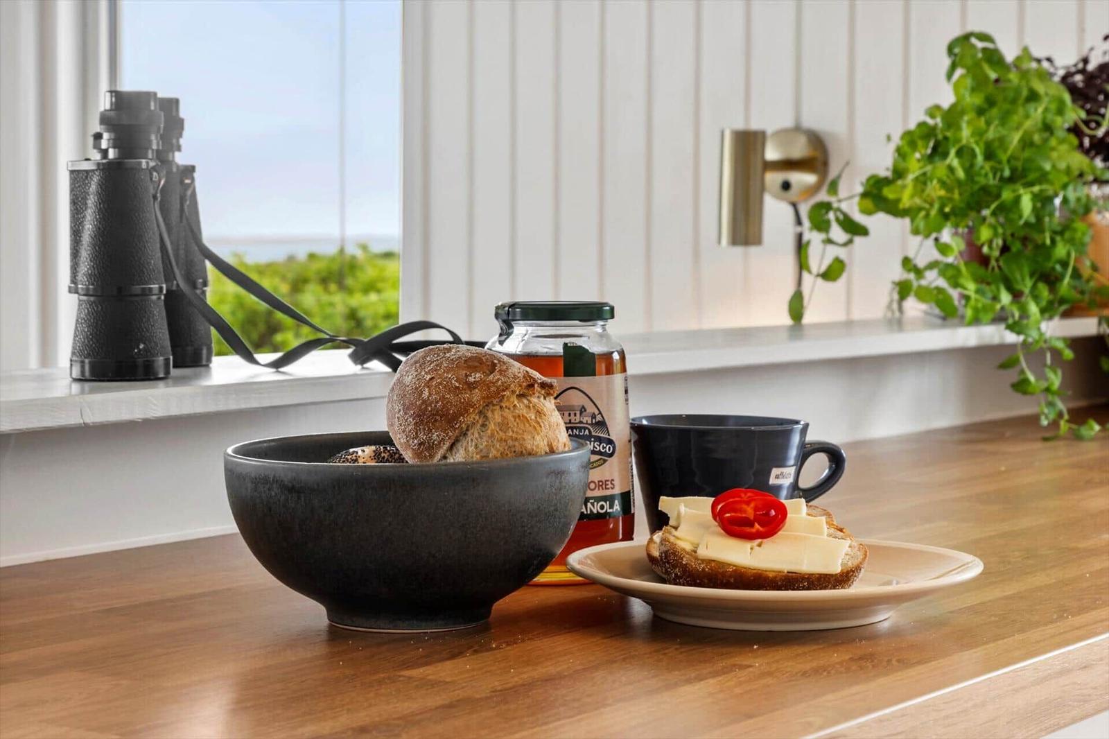 Breakfast with bread, cheese, and tomato on wooden table. Background window with view of greenery and binoculars.