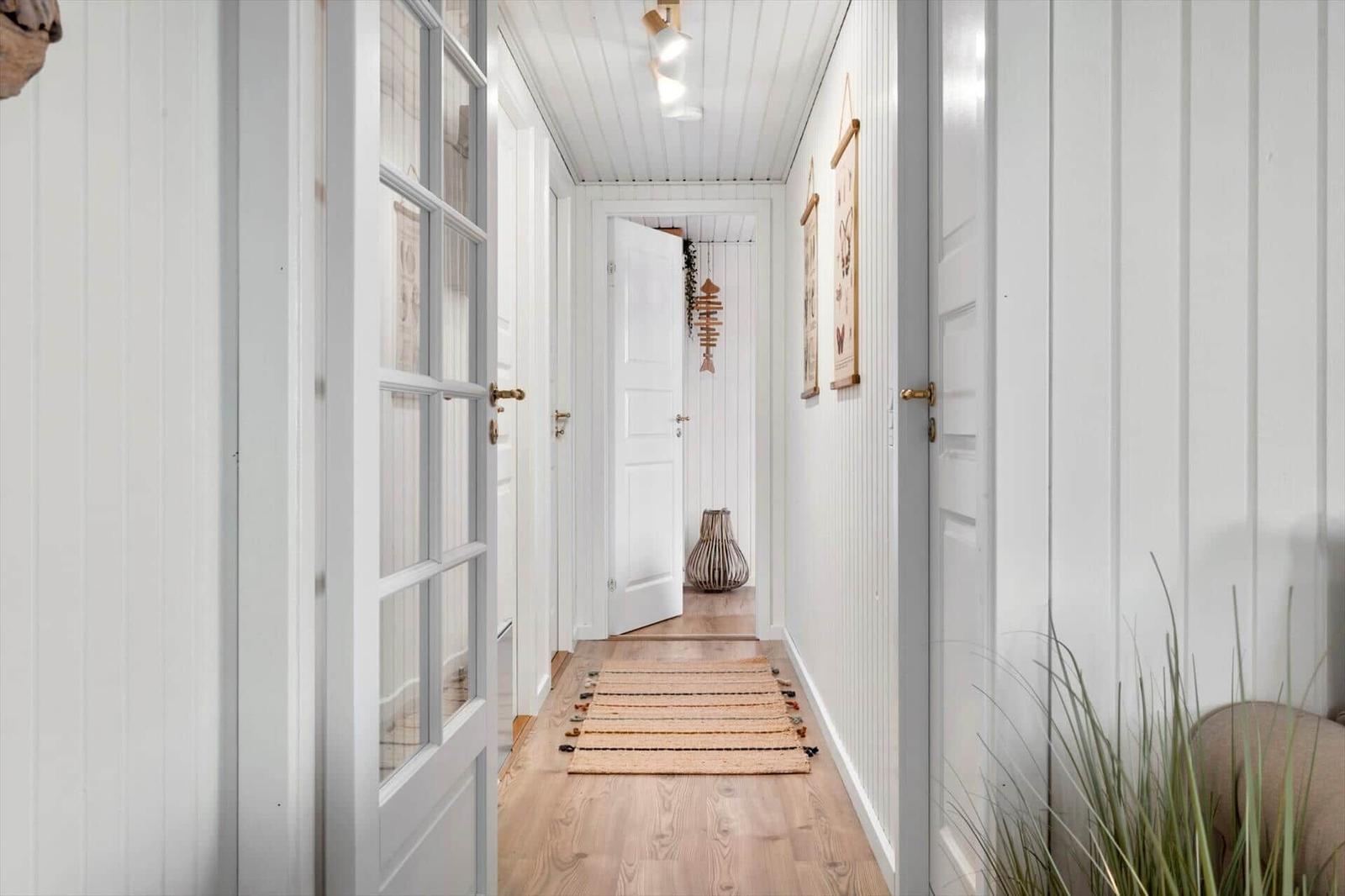Hallway with white walls, wooden floor, and doorways. Rug and plant are visible.