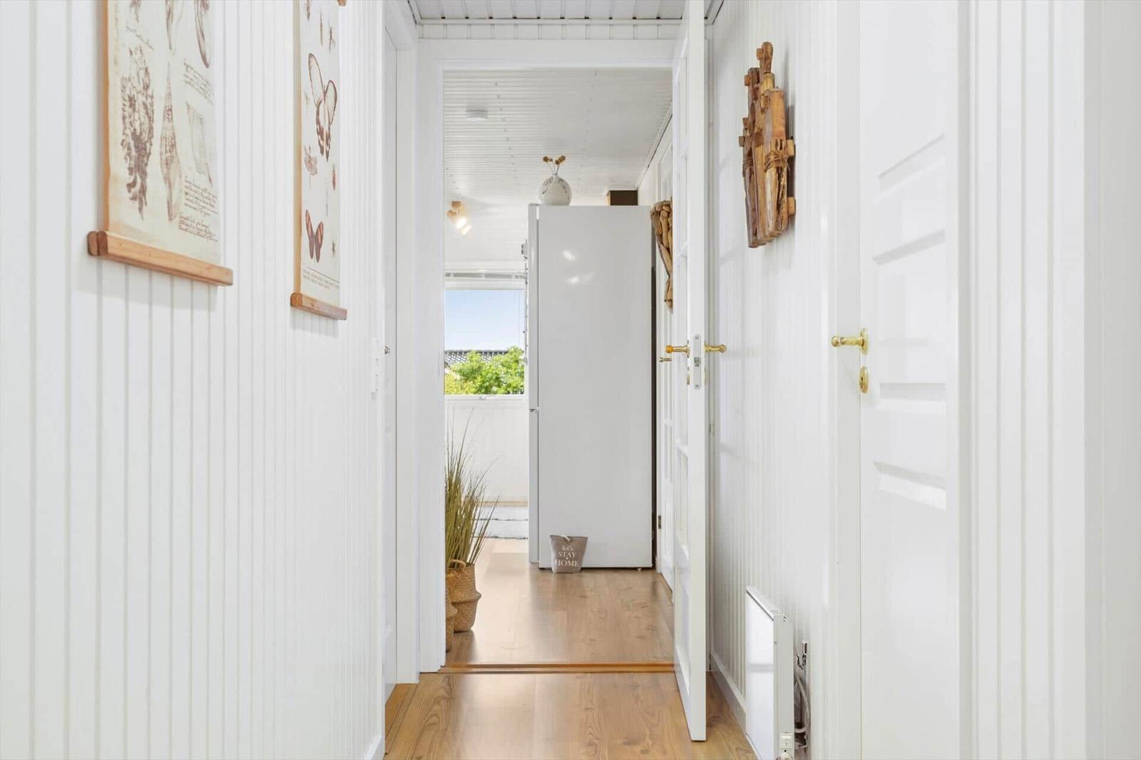 Hallway with white walls, wooden floor, and doors. Pictures and a wooden object hang on the wall.