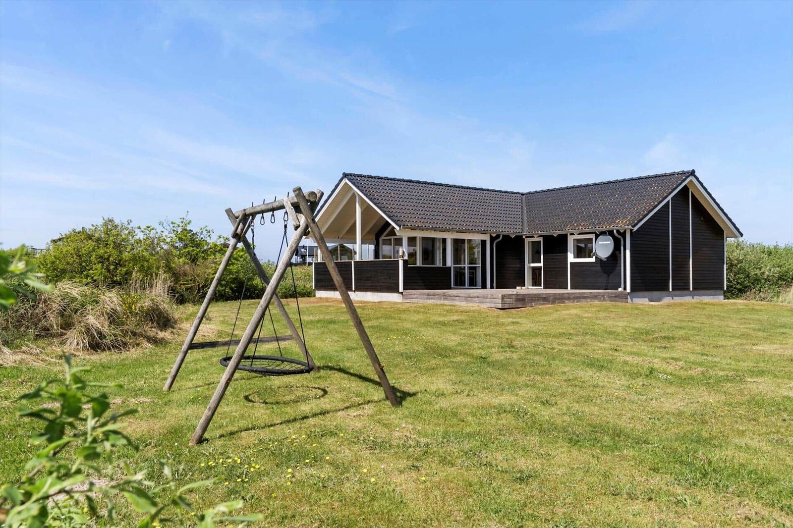 Ein Haus mit dunkler Fassade und Holzveranda. Vor dem Haus steht ein hölzerner Schaukelplatz auf einer Wiese.