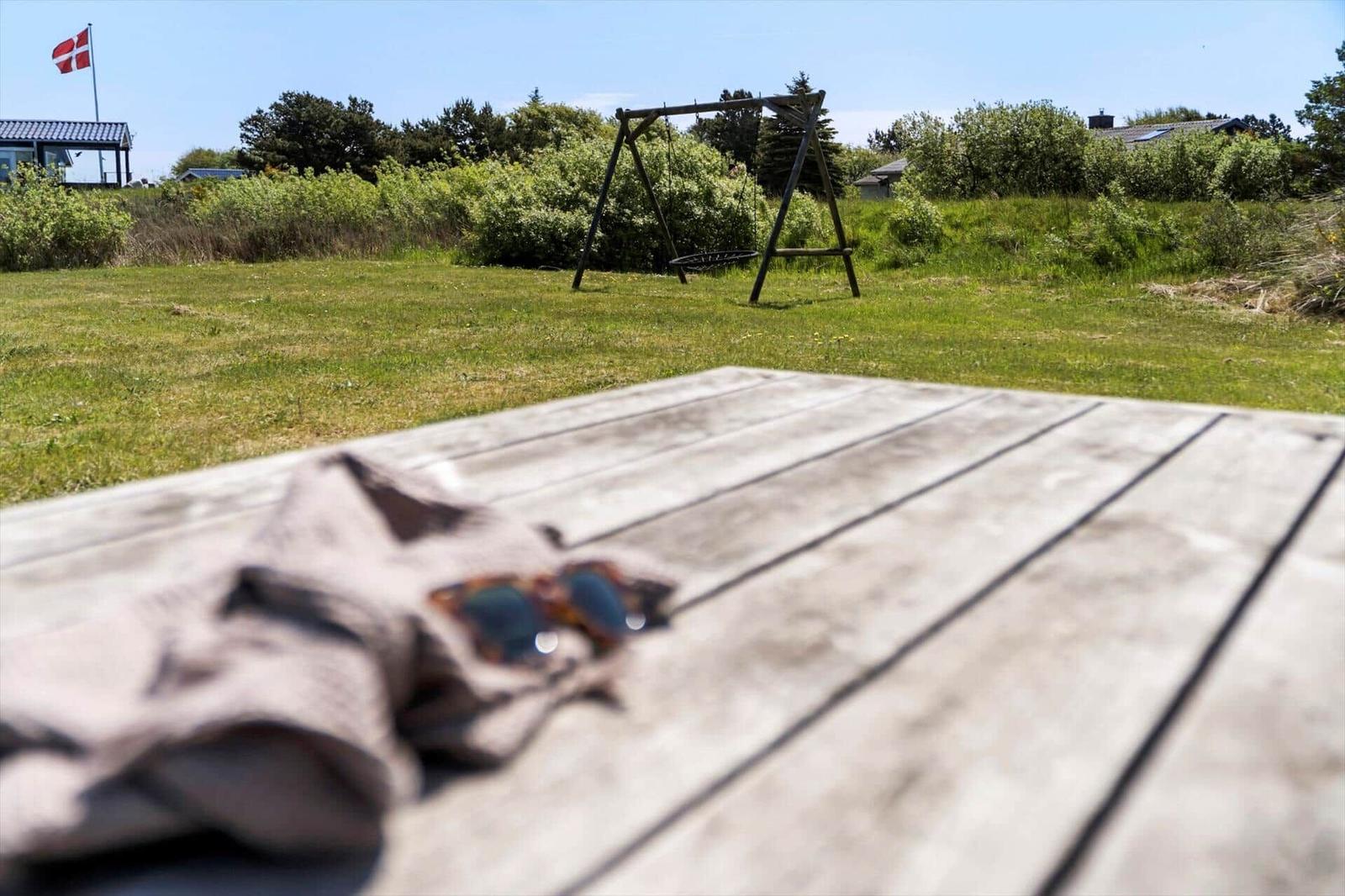 Holztisch mit Sonnenbrille und Tuch im Vordergrund. Hintergrund: Grasfläche mit Schaukel und dänischer Flagge.