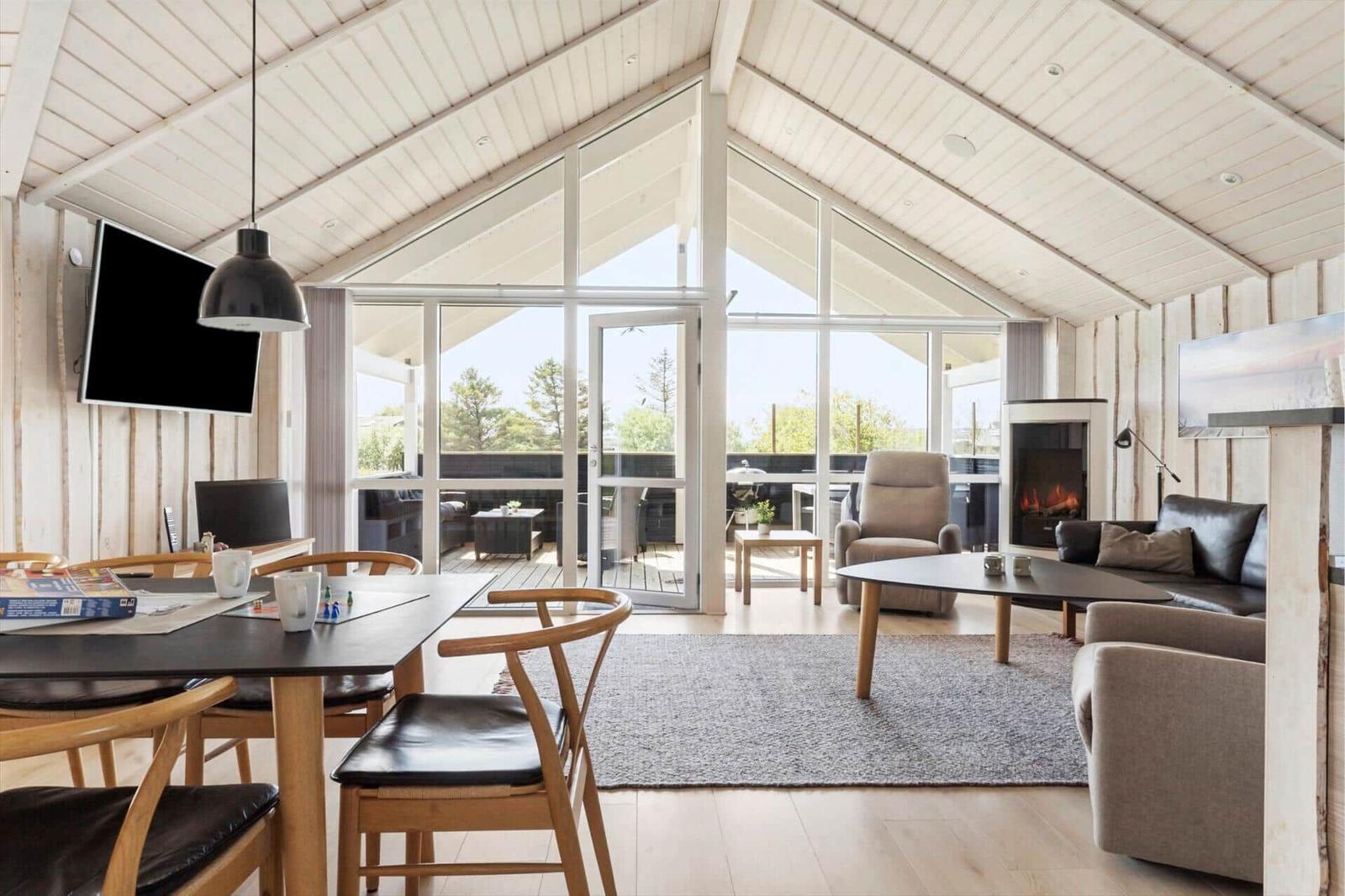 Wohn- und Essbereich mit Kamin, Glasfront und Holzdecke. Möbel sind modern und schlicht.