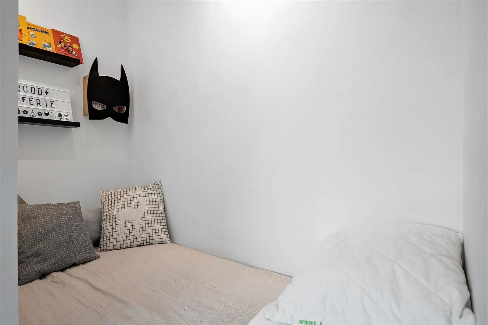 Schlafzimmer mit Bett, Kissen und Wandregal mit Batman-Maske und Büchern.
