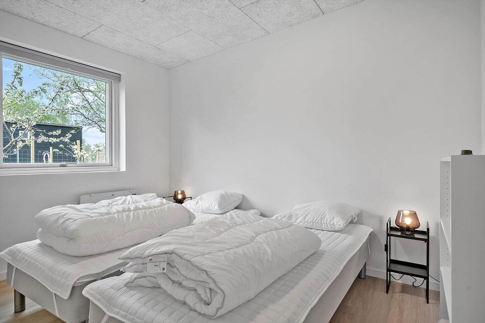Zwei Betten mit weißen Bettwäschen, Nachttisch mit Lampe und Fenster mit Blick auf Bäume.