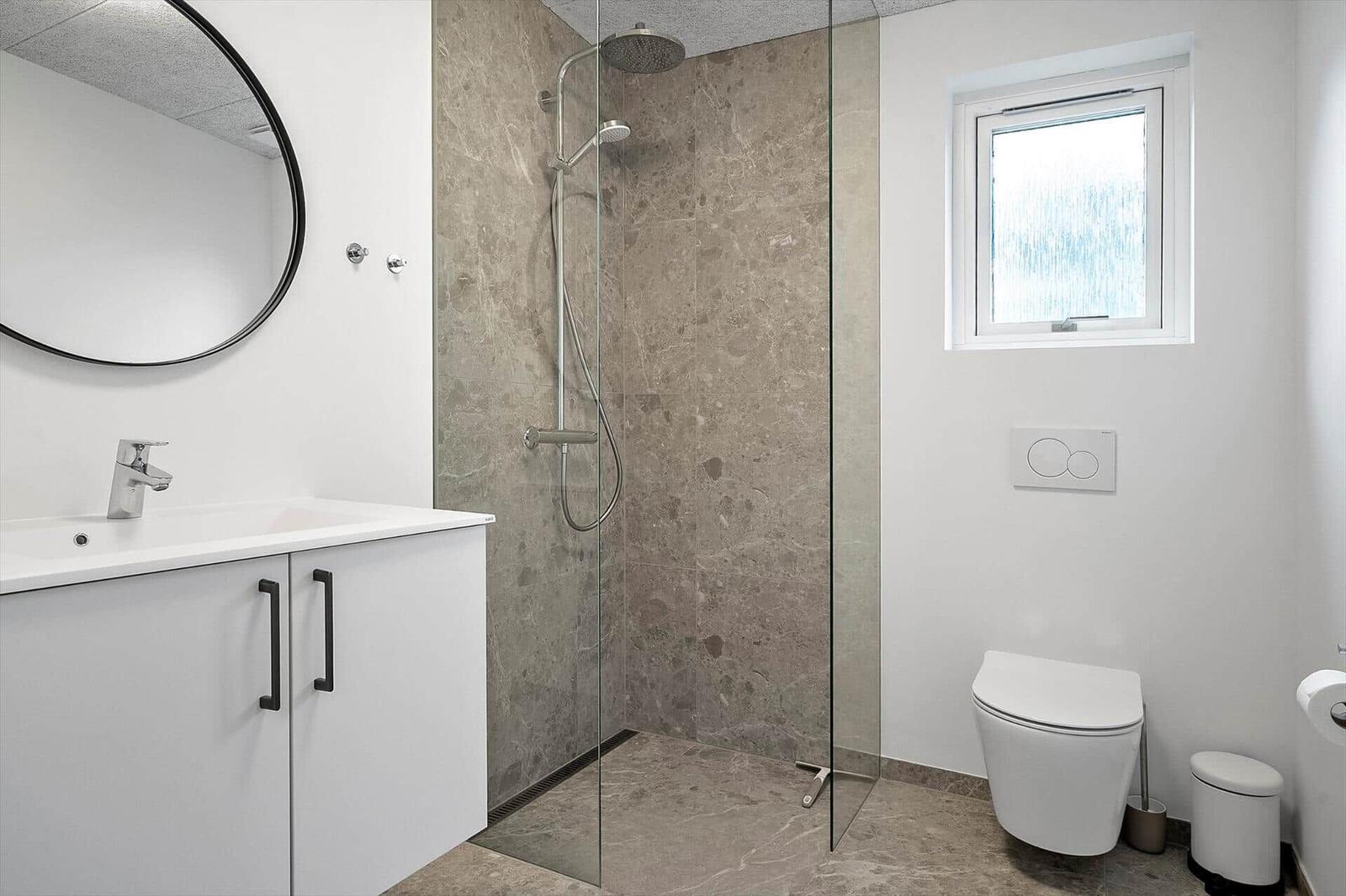 Badezimmer mit Dusche, Waschbecken und Toilette. Glaswand, Marmorfliesen und runder Spiegel.