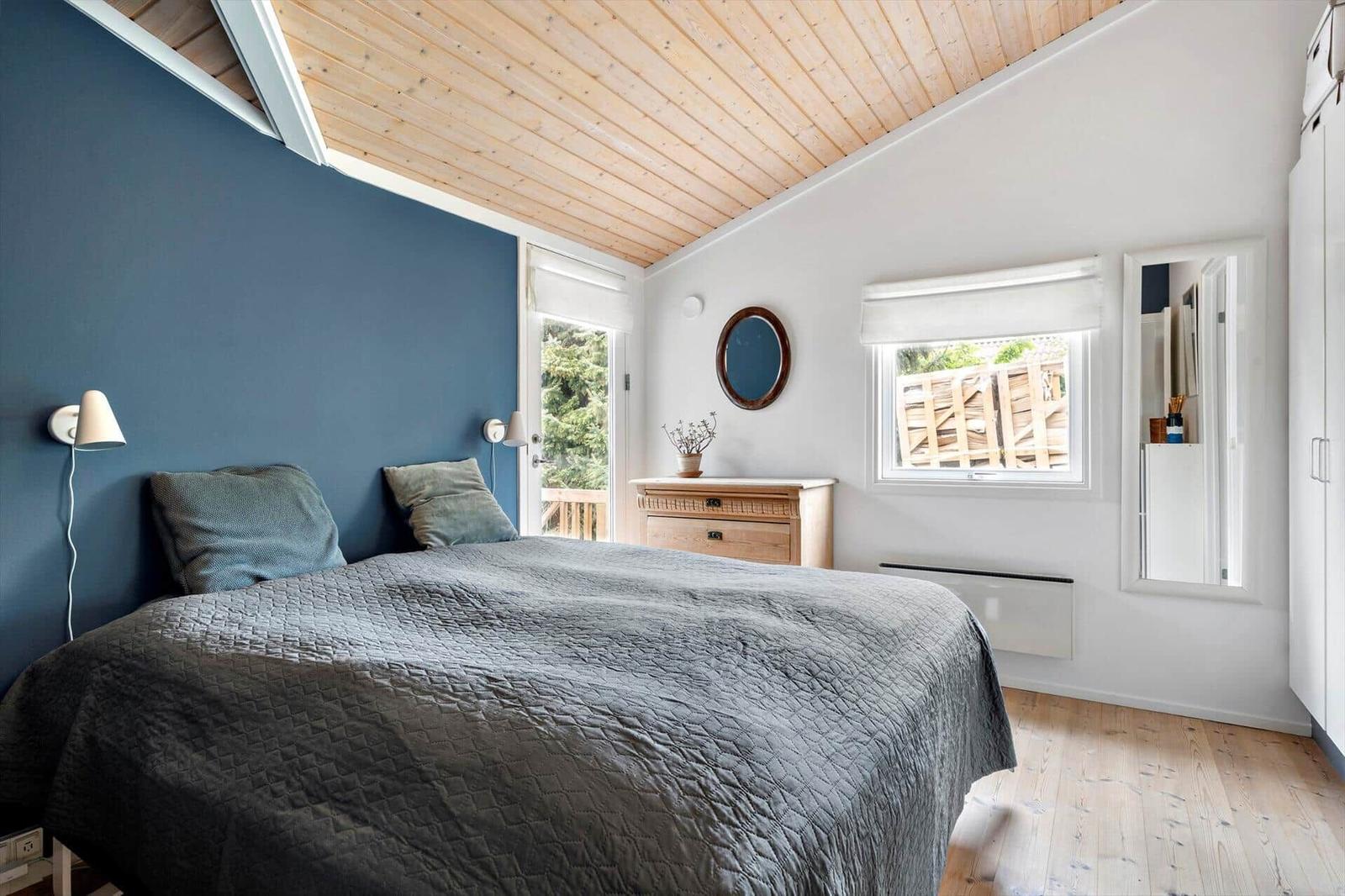 Schlafzimmer mit Bett, Holzdecke und Fenstern. An der Wand hängt ein runder Spiegel.