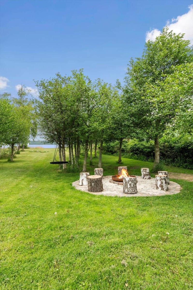 Runder Feuerplatz mit Holzstümpfen umgeben von Gras und Bäumen. Im Hintergrund ist ein See sichtbar.