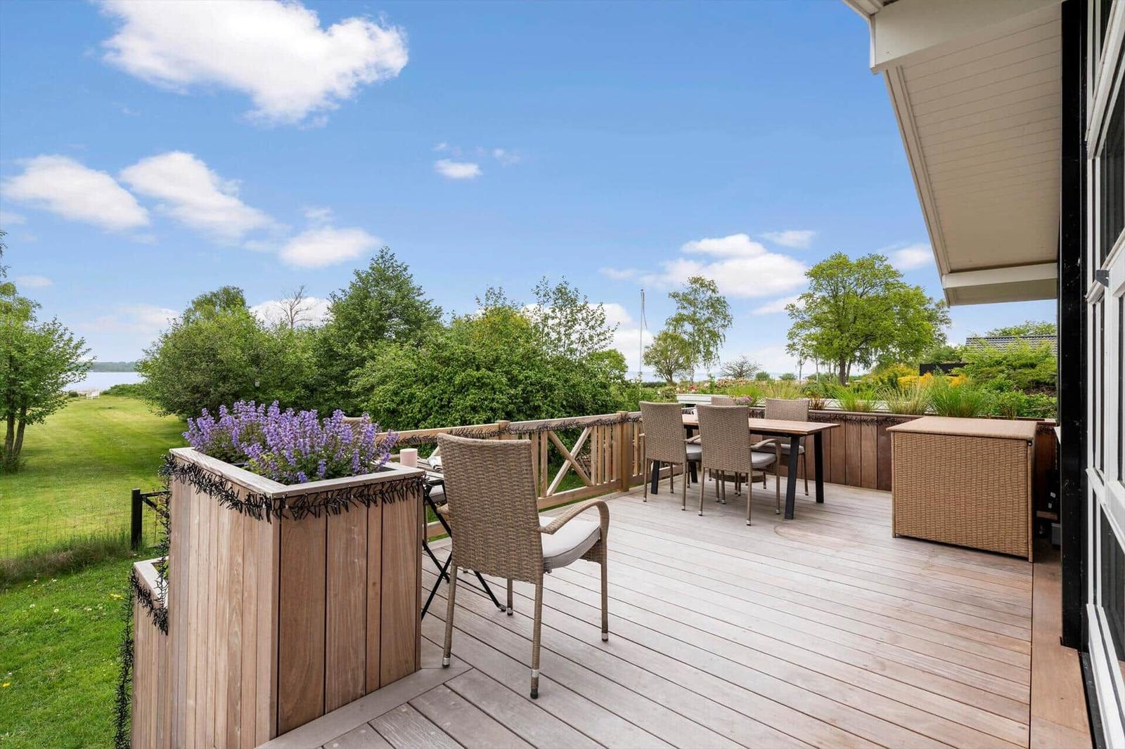 Terrasse mit Sitzgruppe, Holzplanke und Blumenkasten. Blick auf Grünflächen und Bäume.