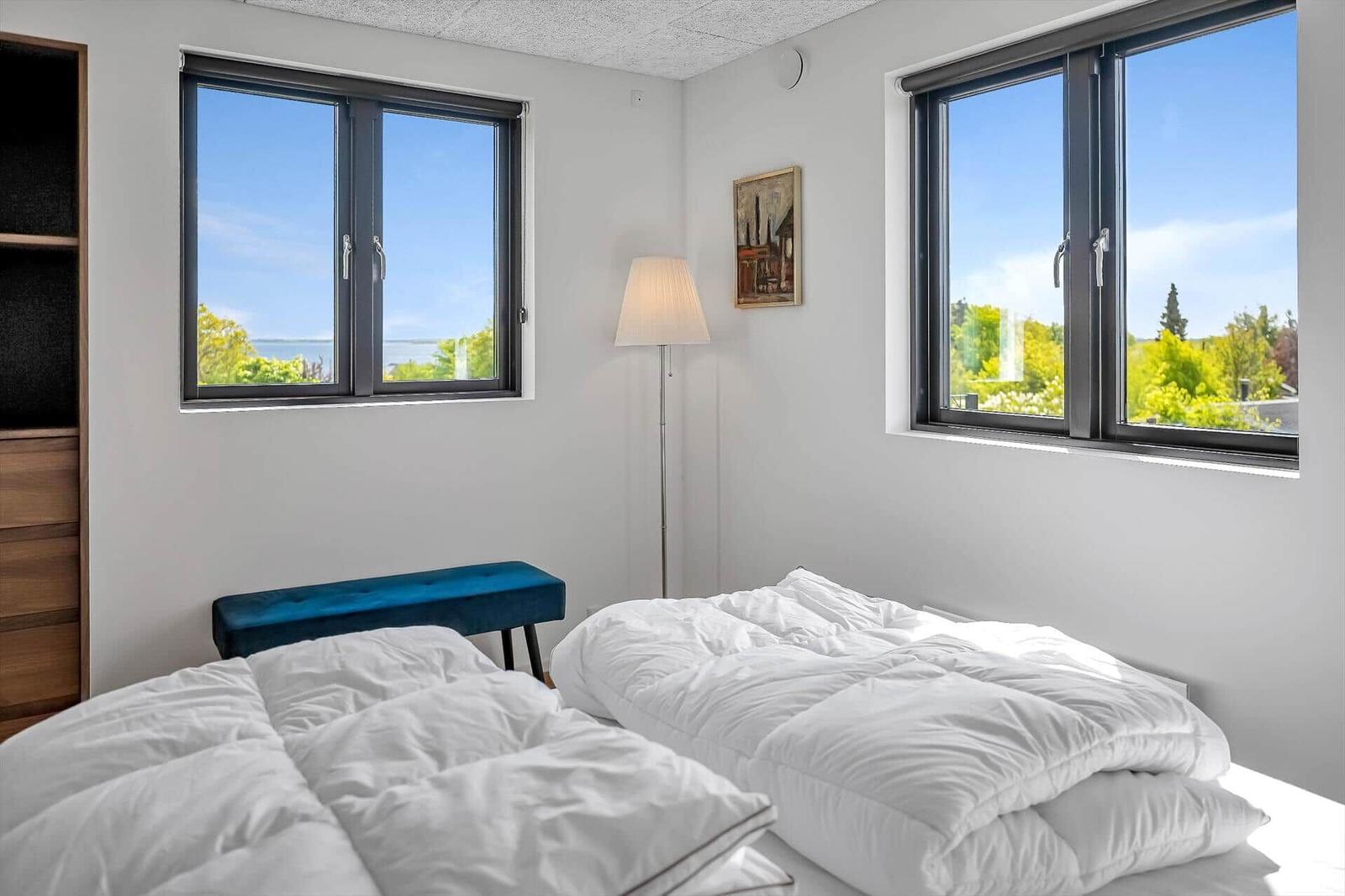 Room with two windows, view of water and greenery, bed with white linens, blue bench.