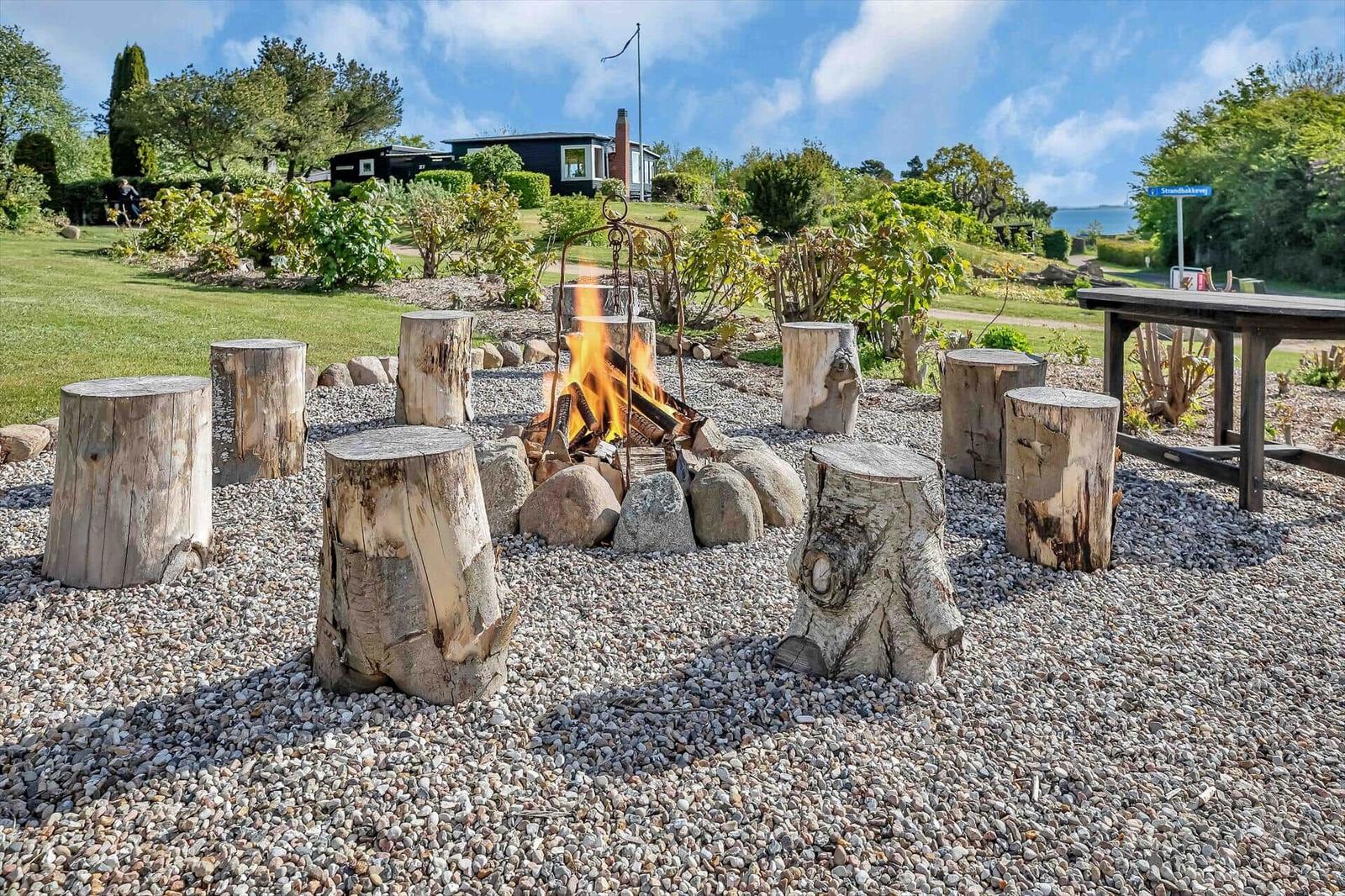 Fire pit with log stumps on gravel. House and greenery in the background.