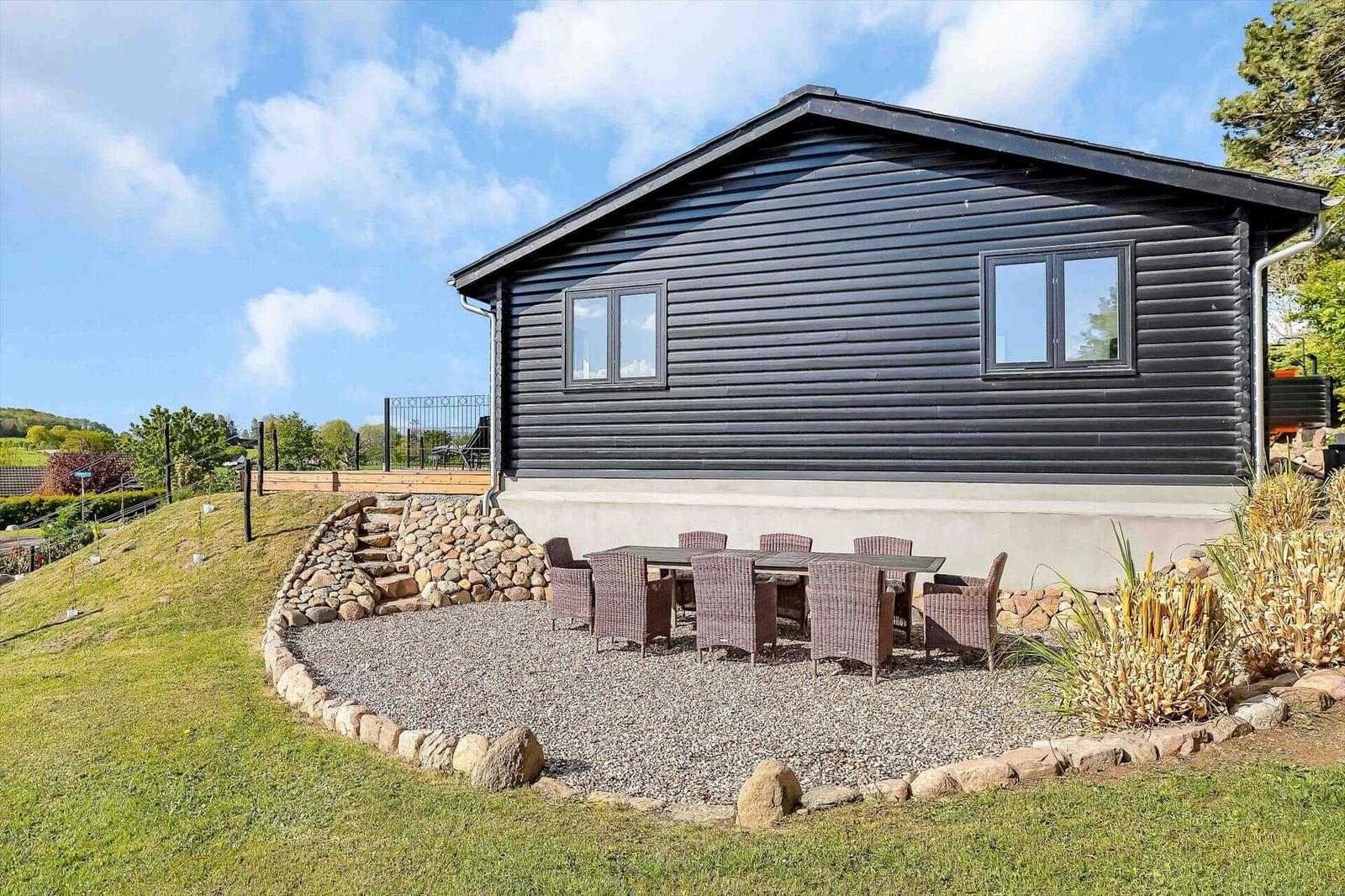 Black wooden house with outdoor seating area. Grassy area and stone walls surround the terrace.
