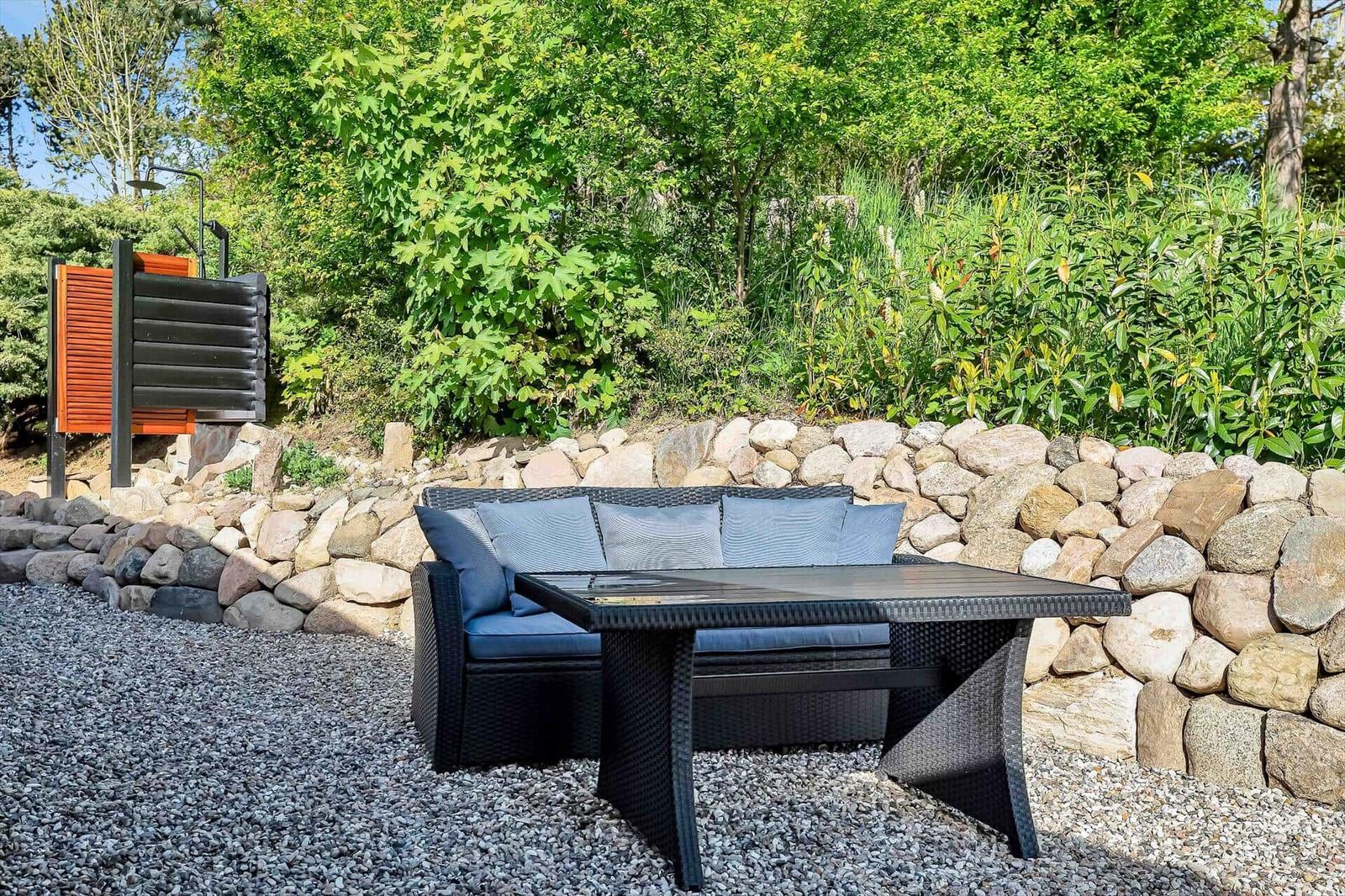 Outdoor area with seating, table, and stone wall. Background: greenery and shower area.