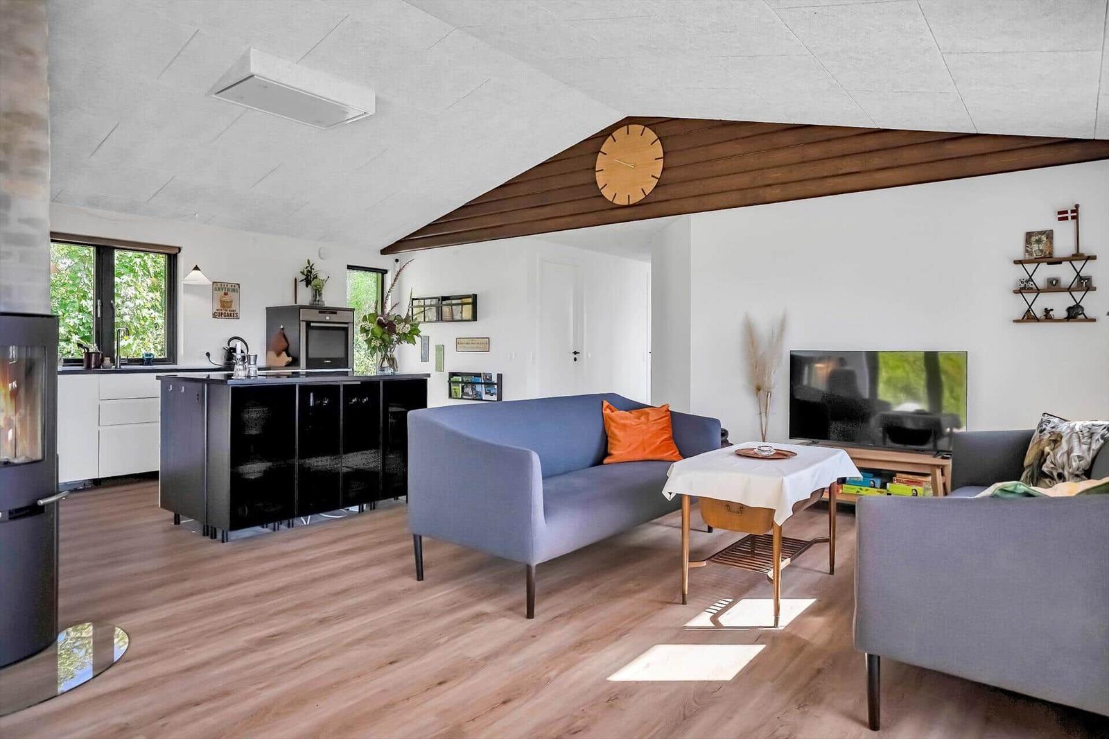 Living area with sofa, table, and TV. Kitchen with countertop and windows.