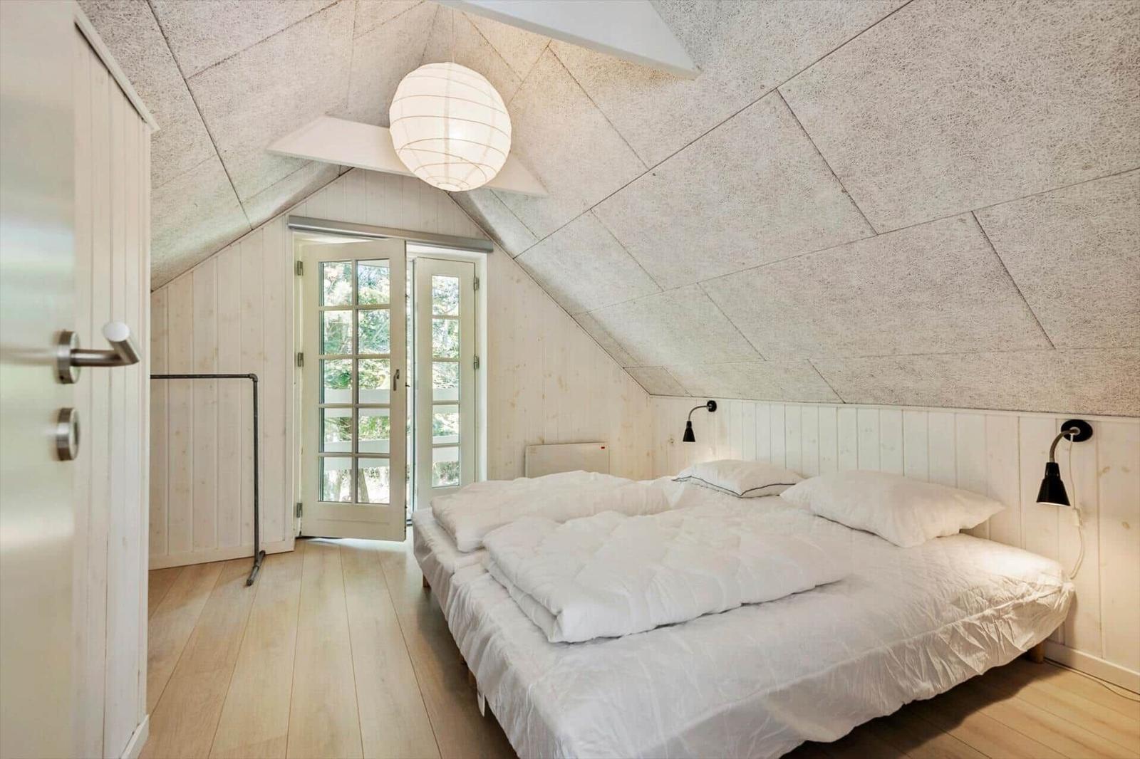 Schlafzimmer mit Dachschrägen, weißem Bett und Holzboden. Tür führt nach draußen.