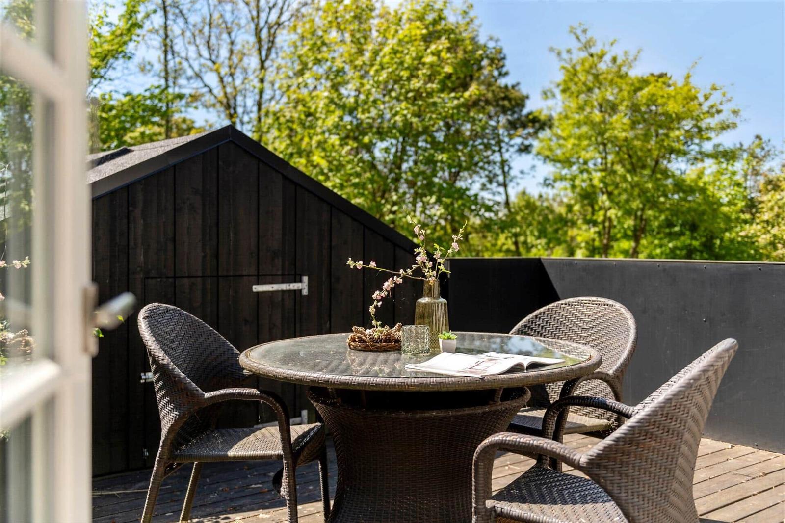 Terrasse mit runden Tisch und Stühlen, dunkler Holzwand und Bäumen im Hintergrund.