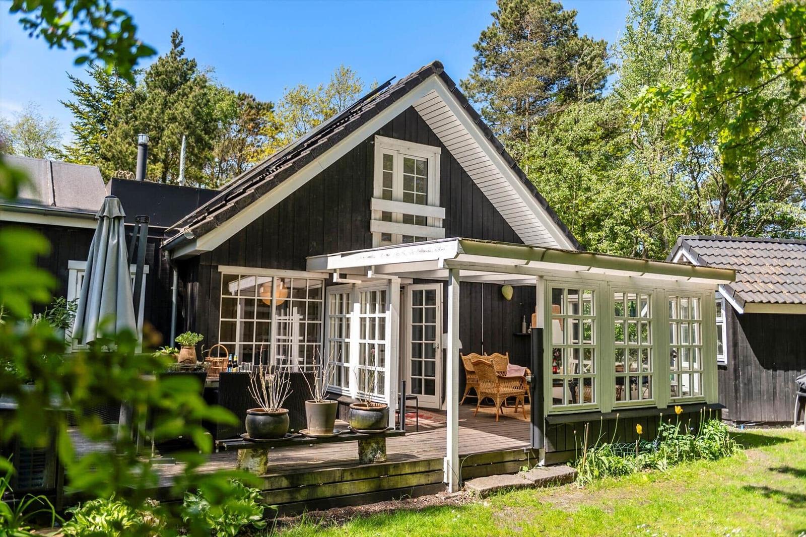 Schwarzes Holzhaus mit Terrasse und Glastür. Umgeben von Bäumen und Gras.