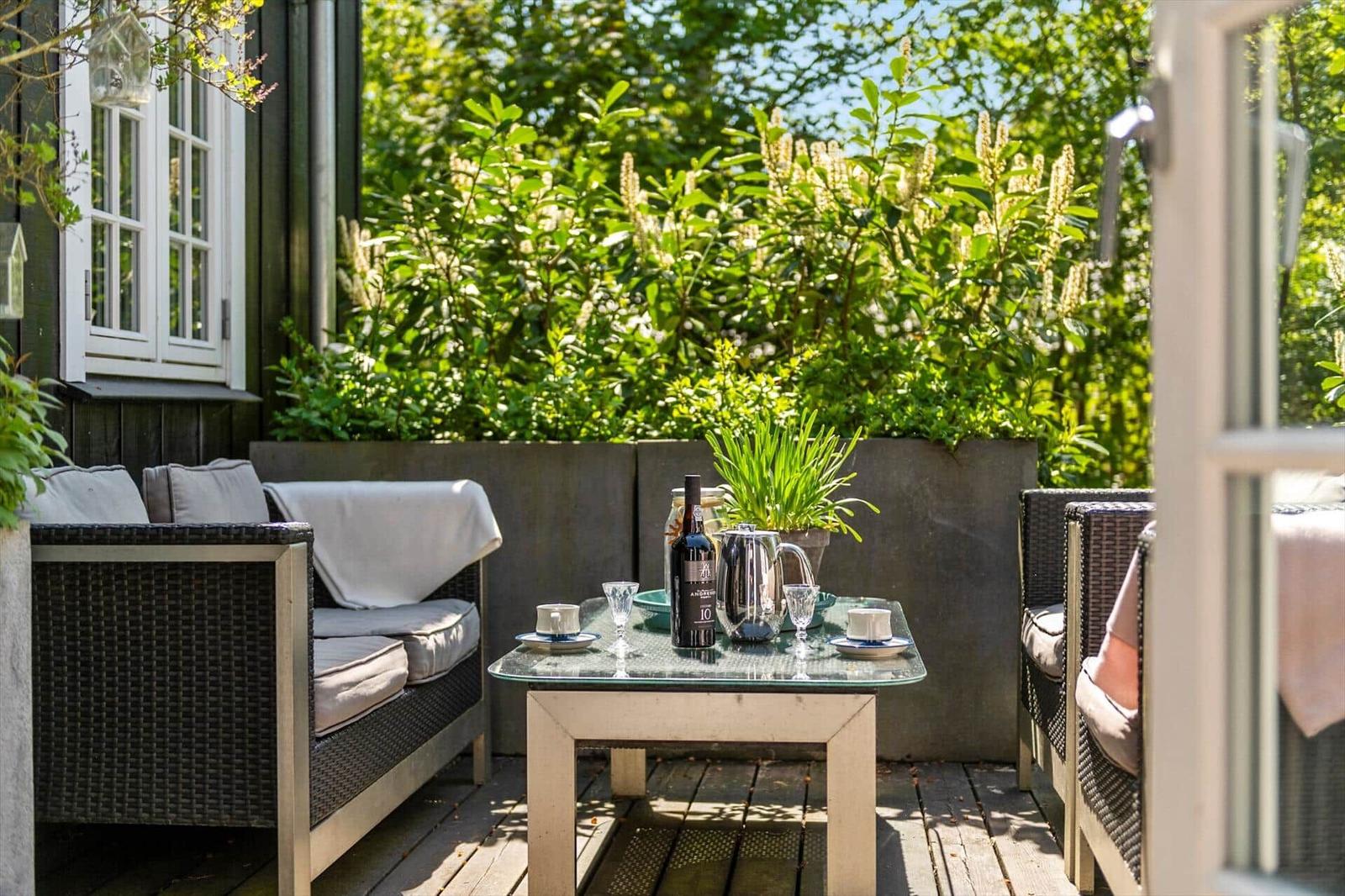 Terrasse mit Sitzgruppe, Tisch und Pflanzen. Hintergrund: grüne Hecke und Fenster.