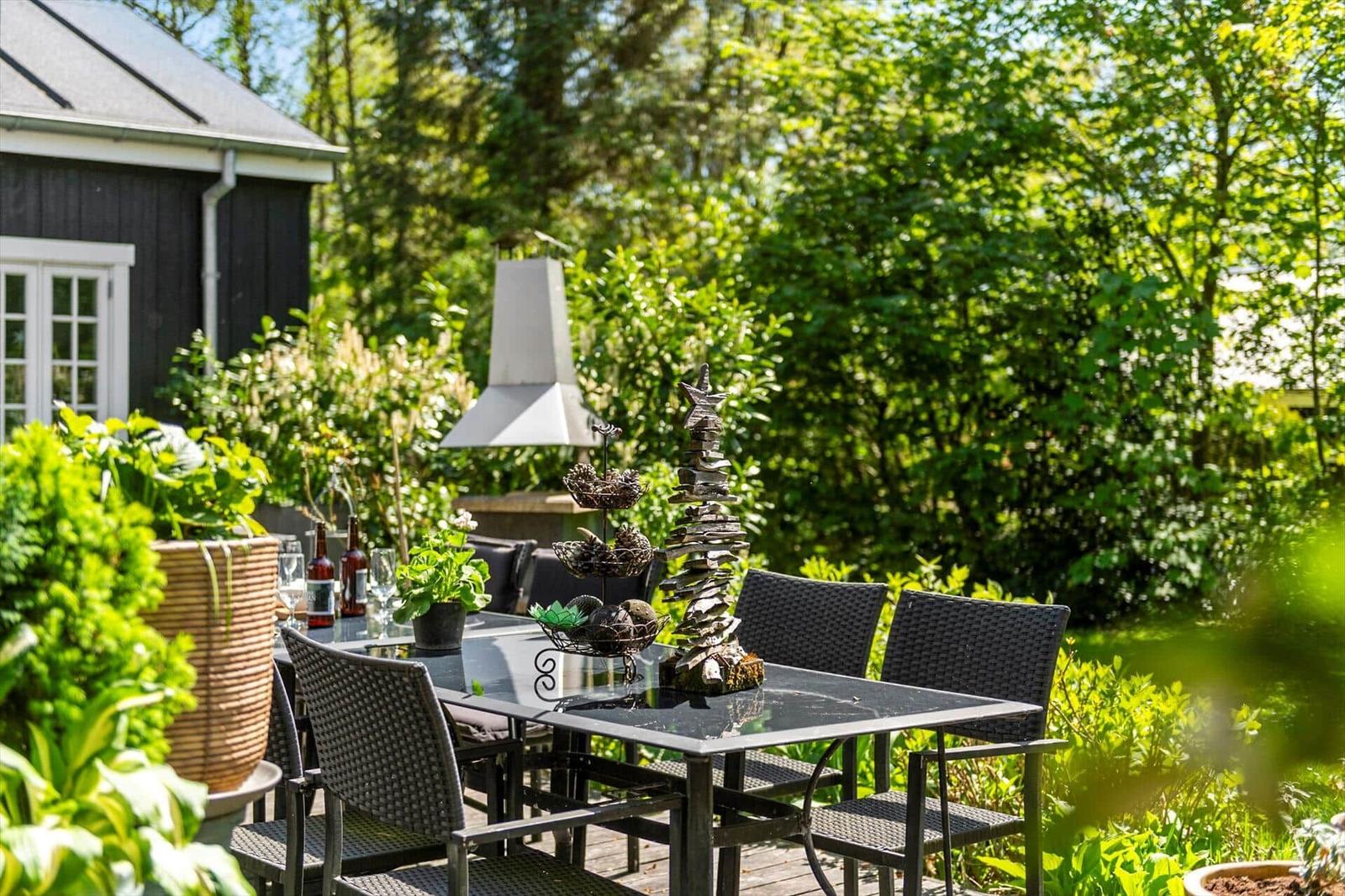 Außenterrasse mit Tisch, Stühlen und Grill, umgeben von grünen Pflanzen und Bäumen.