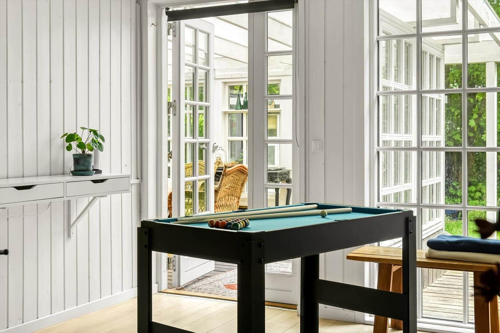 Schwarzer Billardtisch mit grünem Tuch und Kugeln. Weiße Wände, große Fenster und Pflanze.