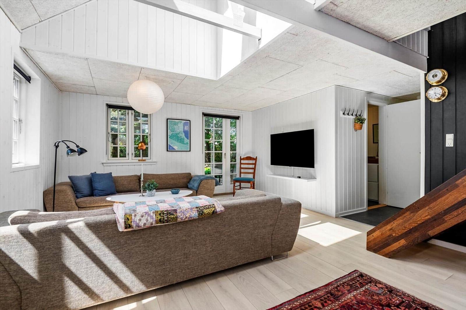 Wohnzimmer mit Sofa, Fernseher und Holzboden. Fenster und Türen führen nach draußen.