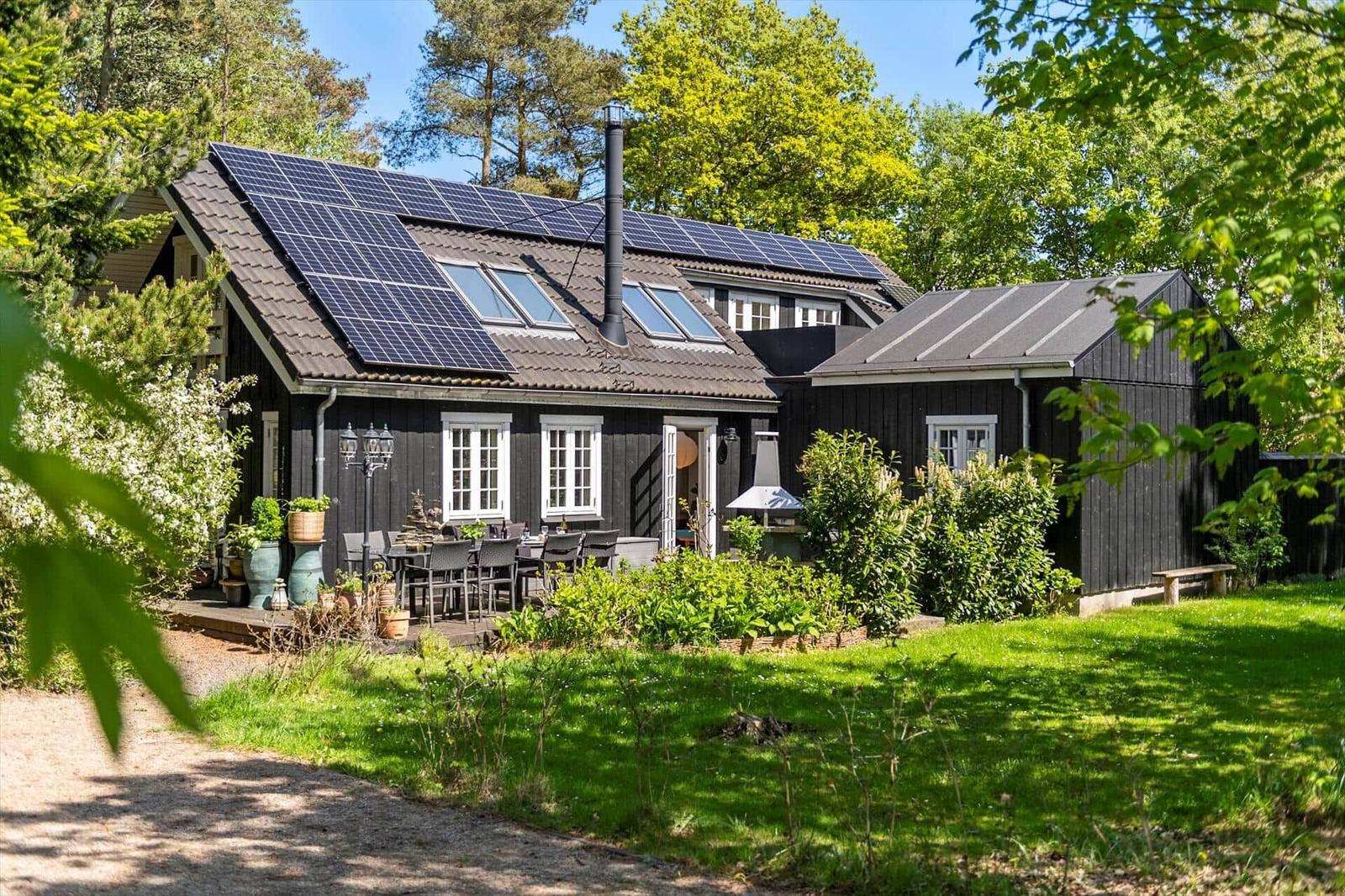 Schwarzes Holzhaus mit Solarpanelen, Terrasse und Garten. Umgeben von Bäumen und Gras.