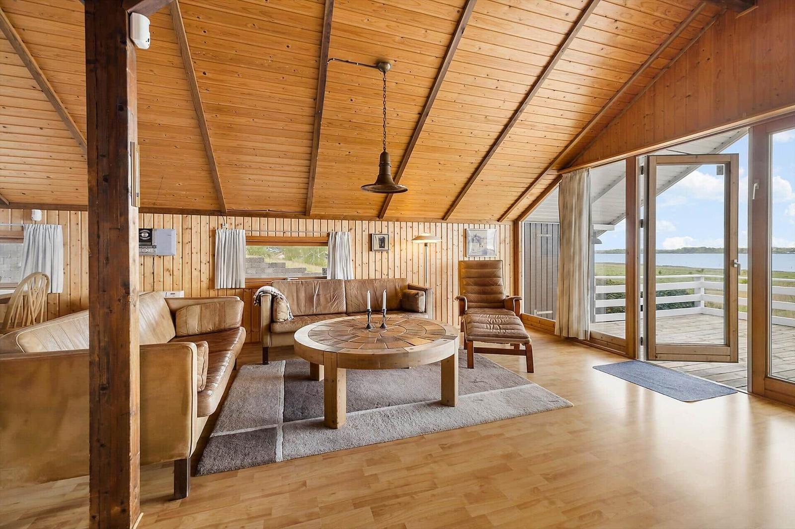Wohnzimmer mit Holzdecke, Ledersofa und Tisch. Durchgang zur Terrasse mit Blick auf Wasser.