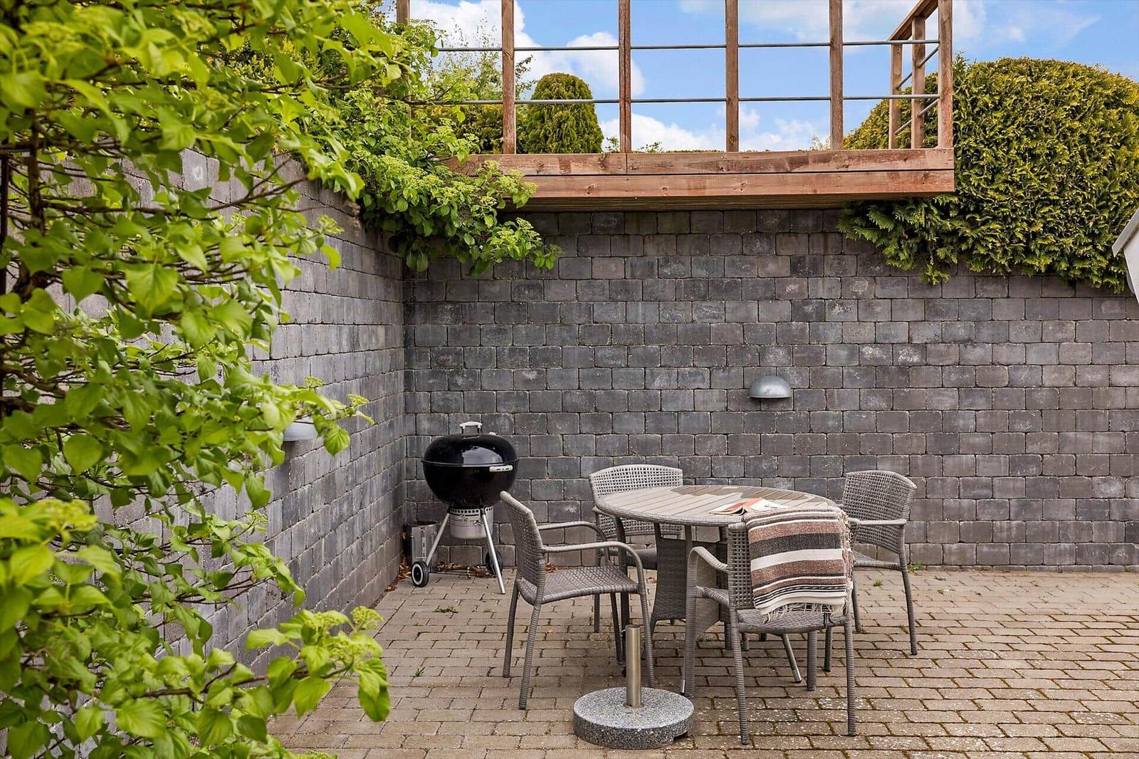 Gartenbereich mit Grill, Tisch und Stühlen. Hintergrund: Steinmauer und Holzterrasse.